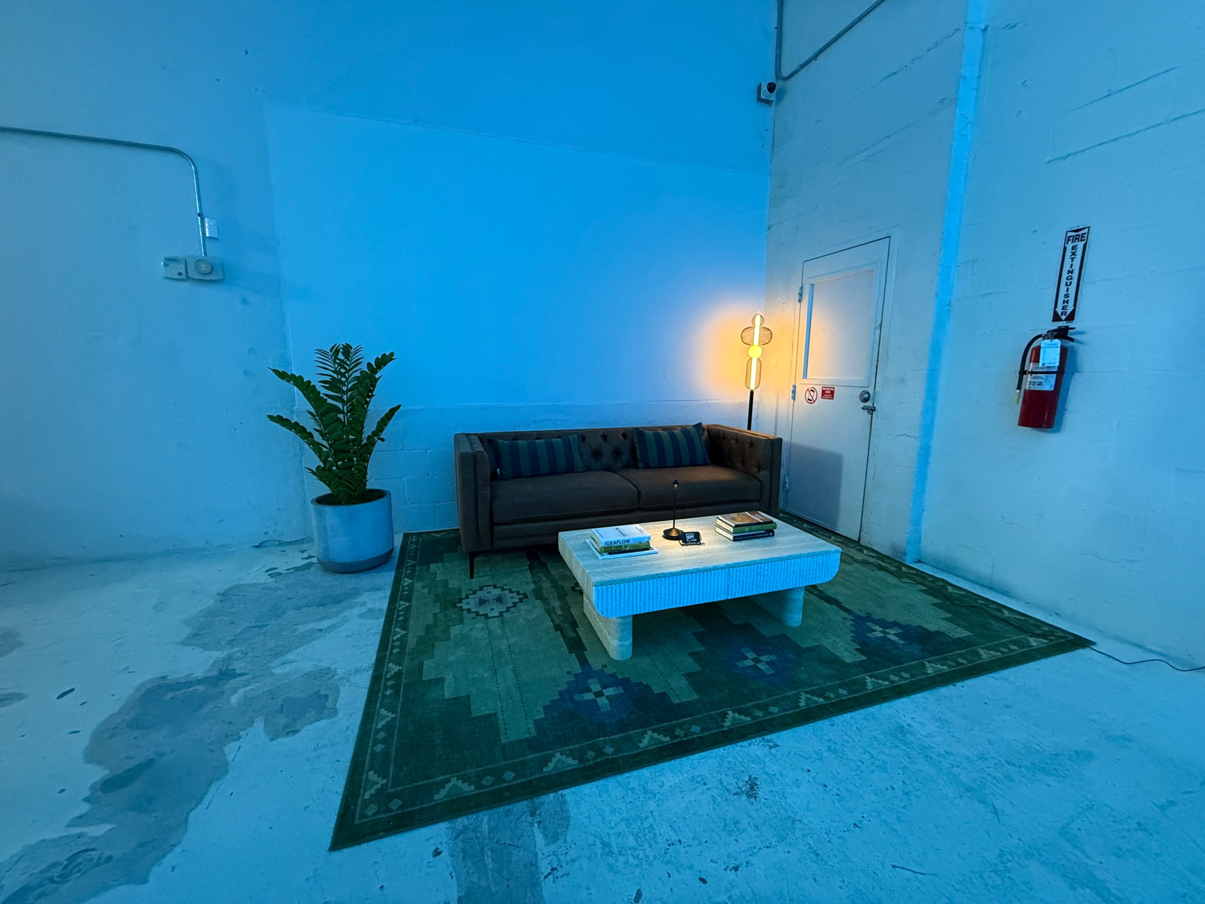 The image shows a minimalistic indoor space featuring a dark-colored couch, a coffee table with books on it, a large potted plant, and a floor lamp, all set against a blue-lit wall.