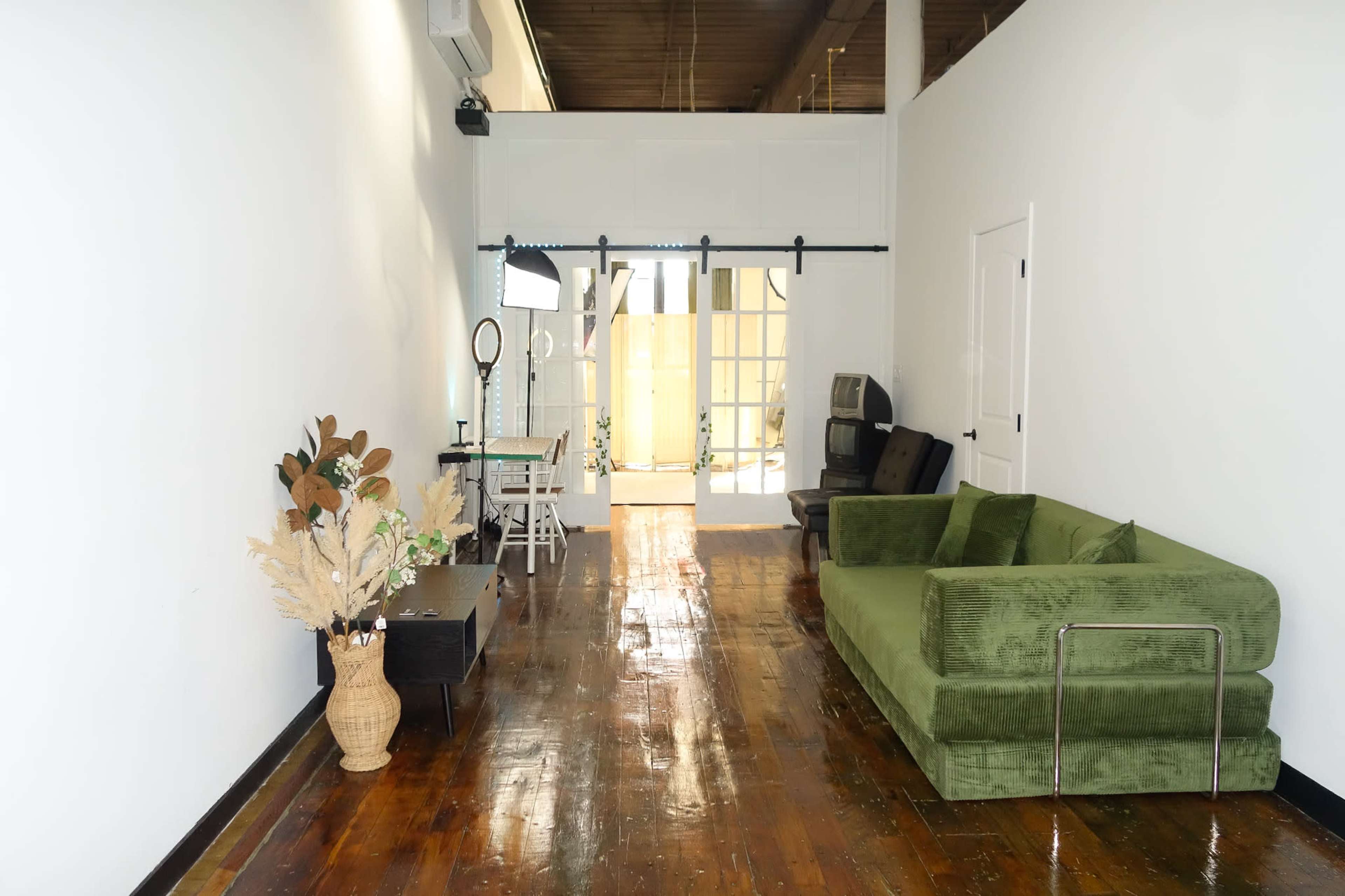 The image shows a spacious, well-lit room with a green sofa on one side, a wooden floor, and a large, bright area beyond a sliding door at the far end.