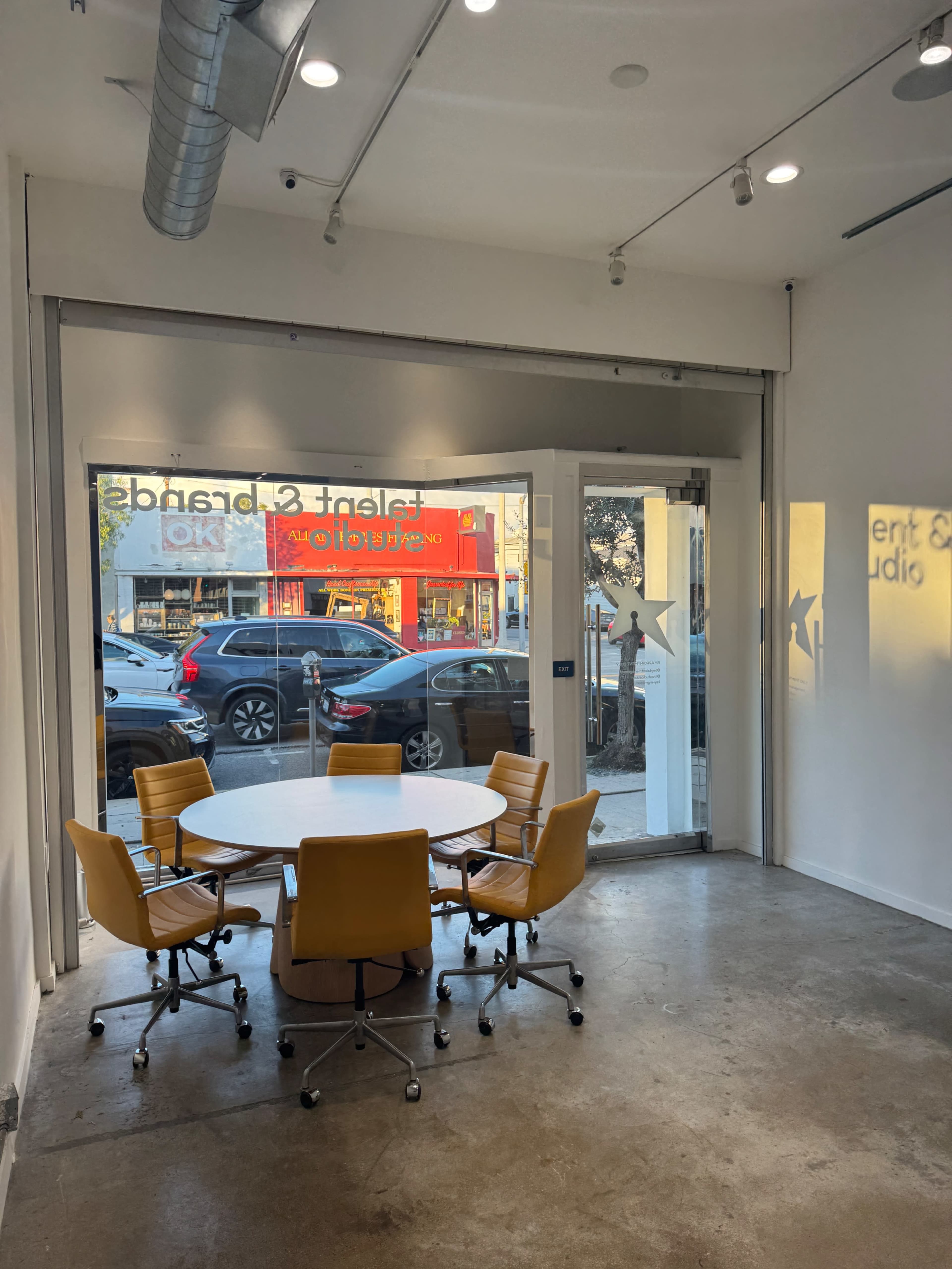 The image shows a modern conference room with a circular table and yellow chairs, featuring large glass doors that open to a view of a street with parked cars and a storefront.