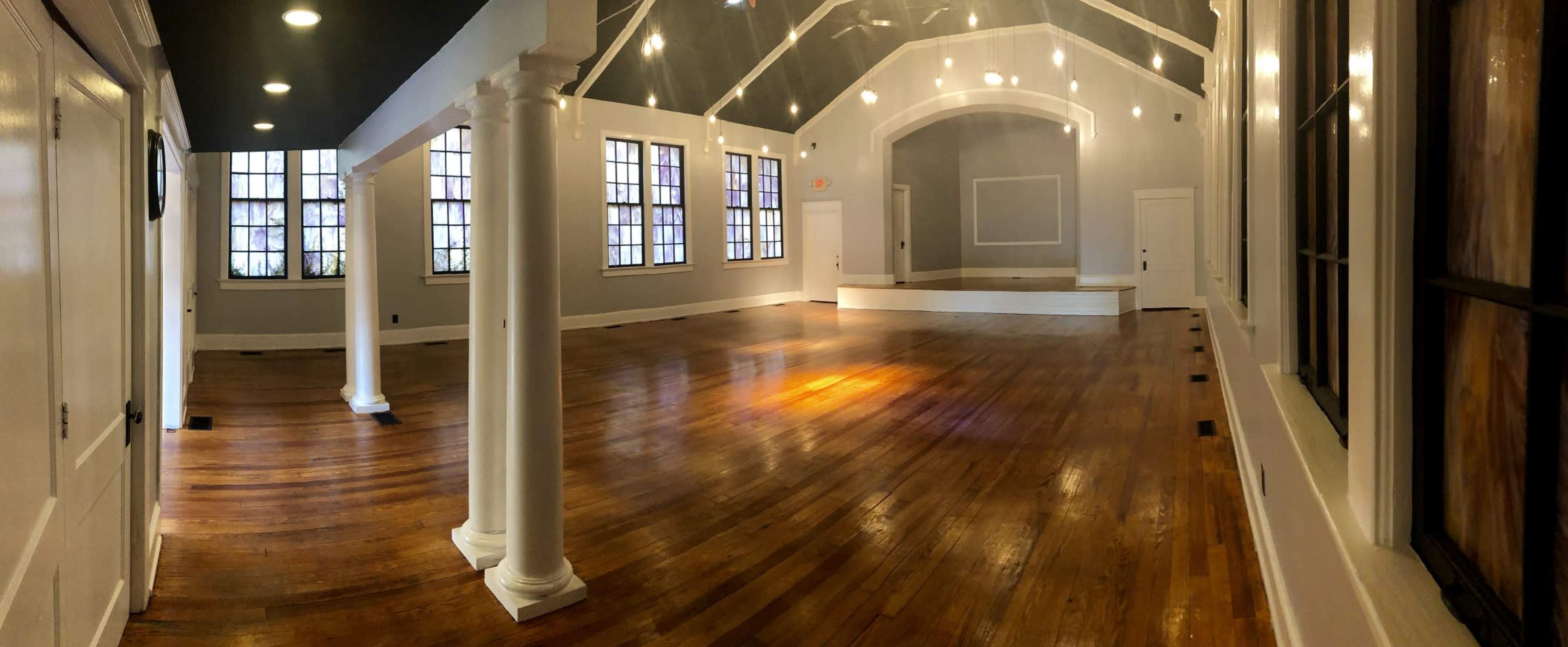 A spacious interior room with wooden floors, white walls, and columns, featuring large windows and a raised stage area at the far end.