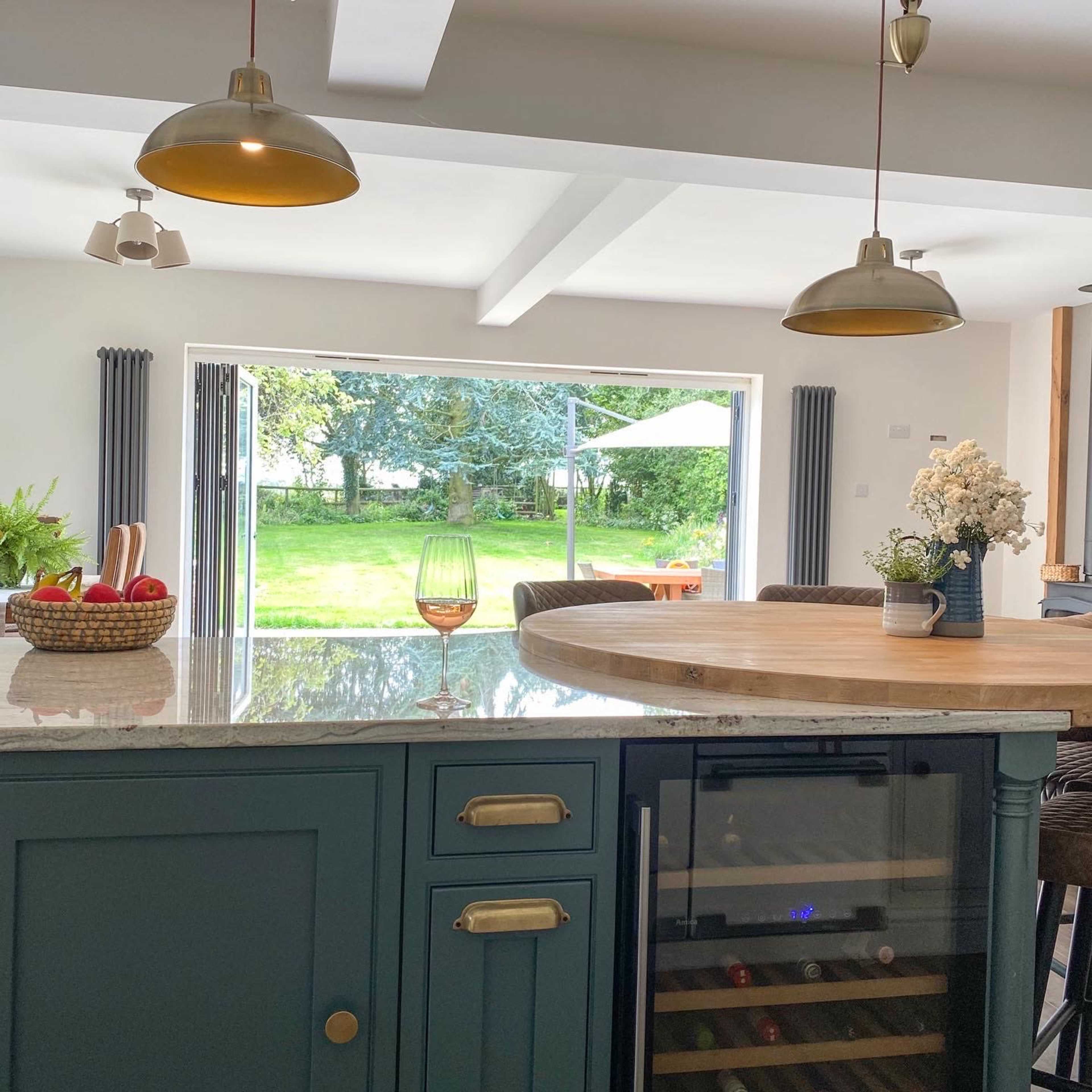 A modern kitchen features a central island with a basket of apples and a wine cooler, opening up to a garden view through large sliding doors.
