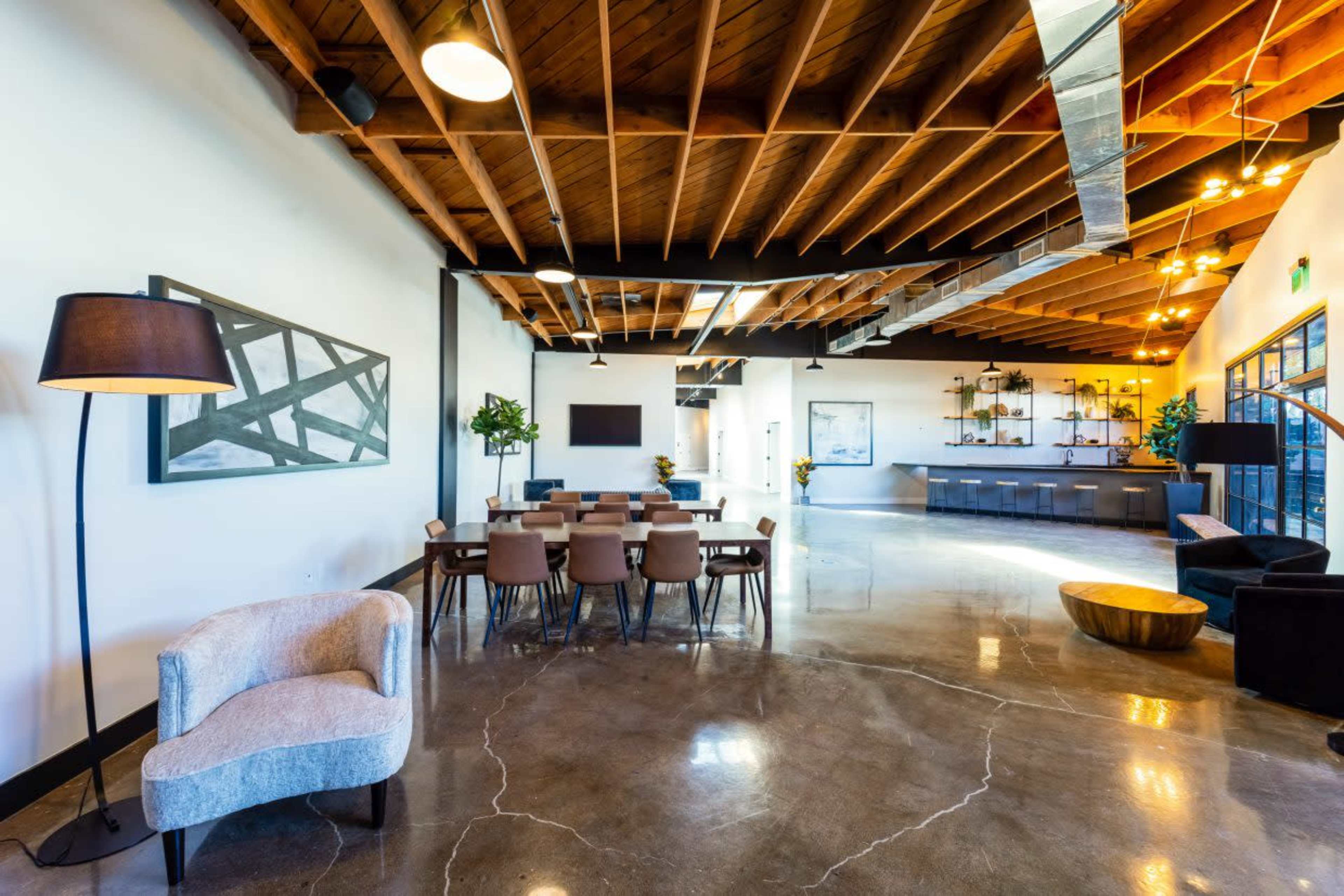 The image shows a modern interior space featuring a dining area with wooden chairs and a table, a seating area with a sofa, and a kitchenette or service area in the background, all under a wood-beamed ceiling.