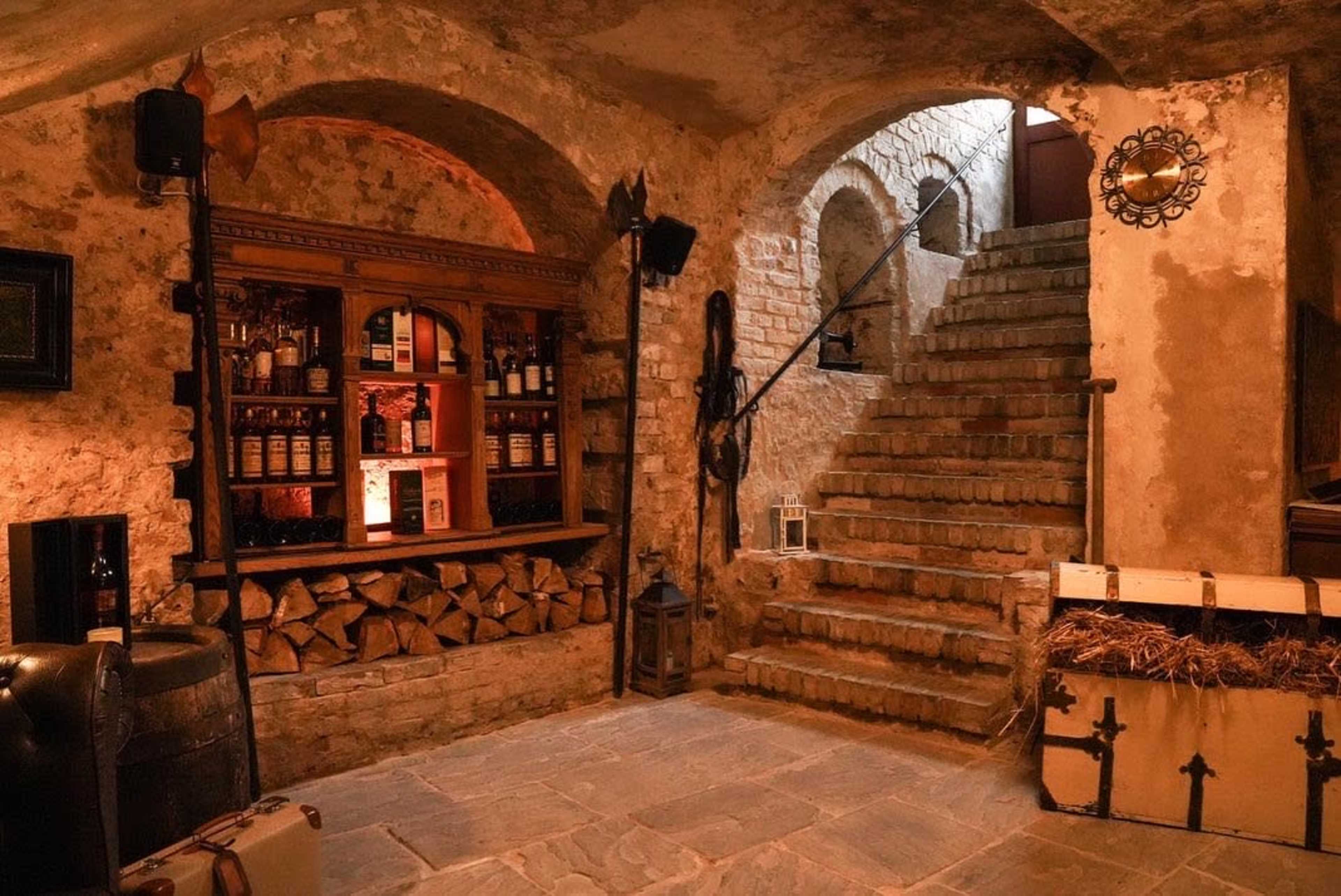 The scene depicts a rustic basement bar area with a wooden shelf stocked with bottles, a stack of firewood, and a staircase leading to a higher level.