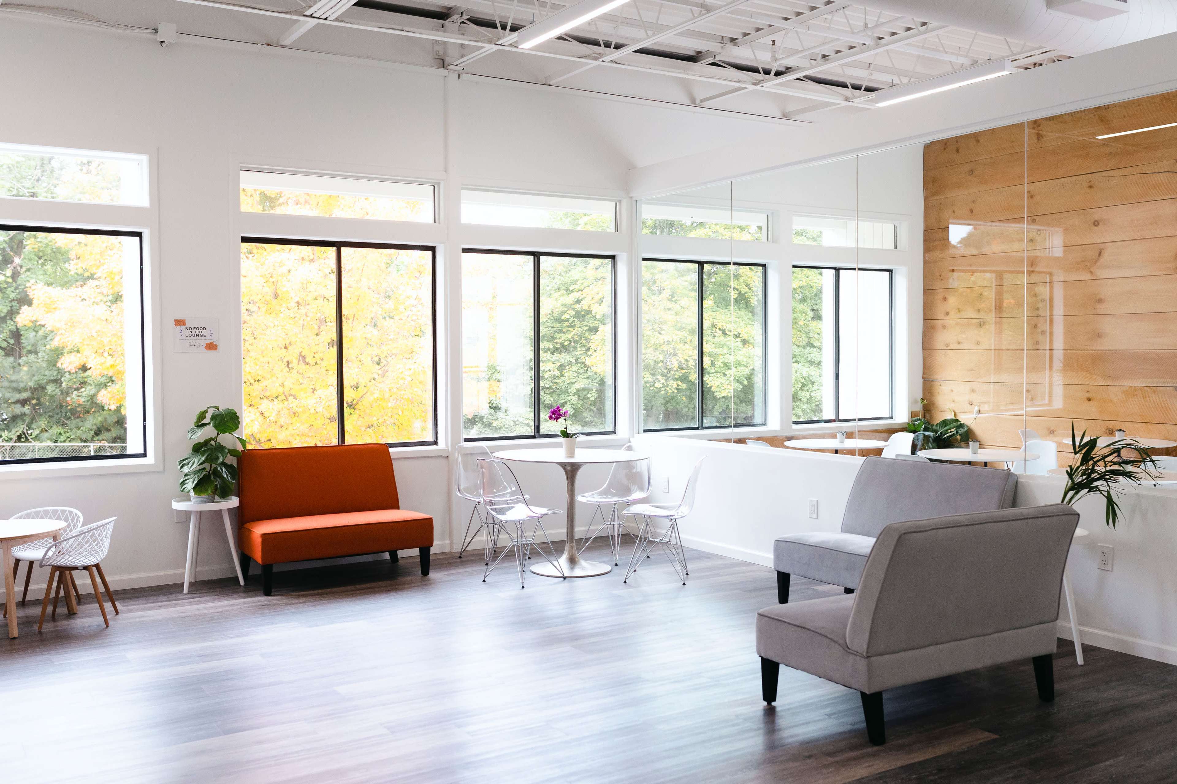 A bright, modern office space with large windows, a gray sofa, an orange chair, and a small table with clear chairs.