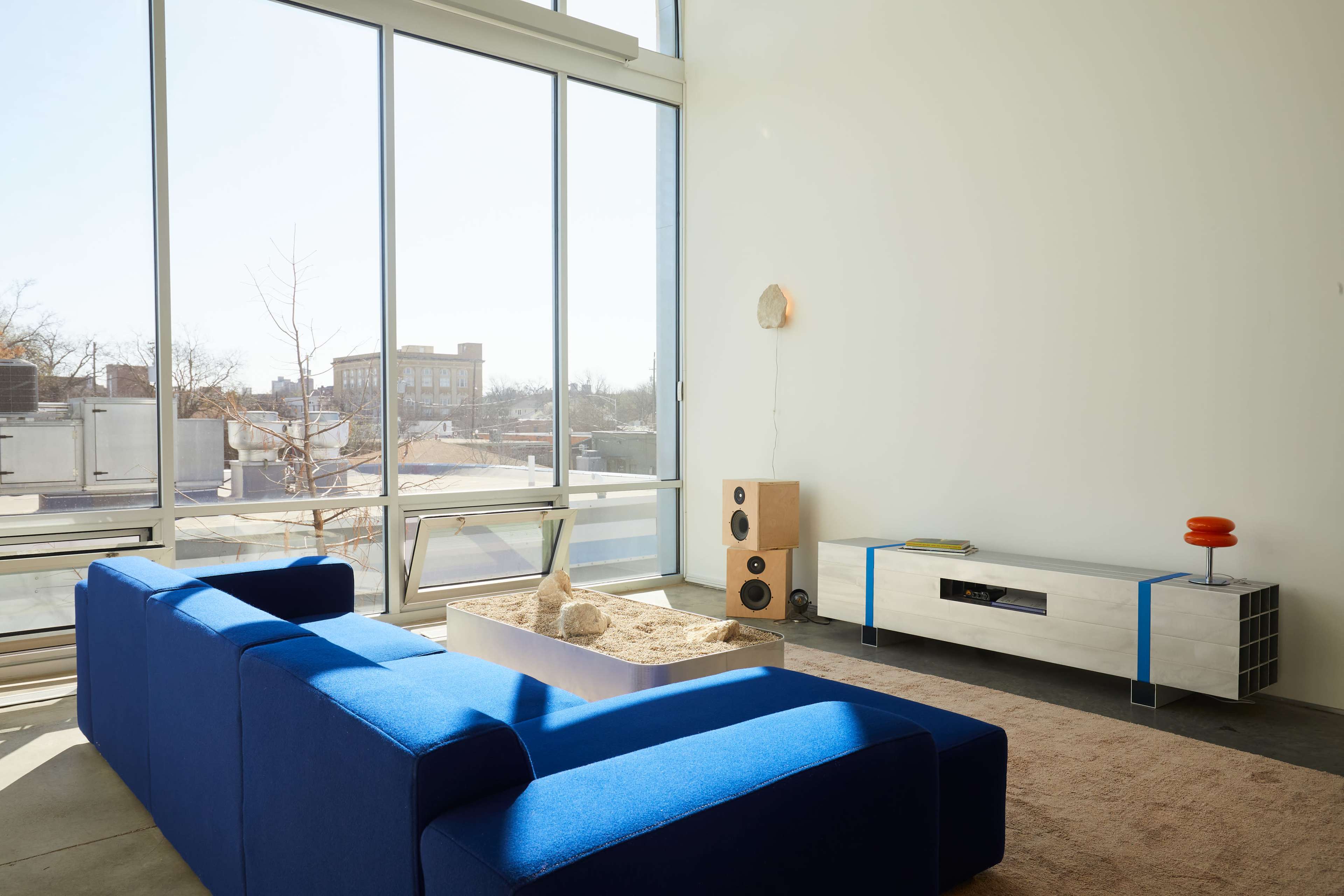 A blue sectional sofa faces a modern media console in a bright living room with large windows overlooking a cityscape.