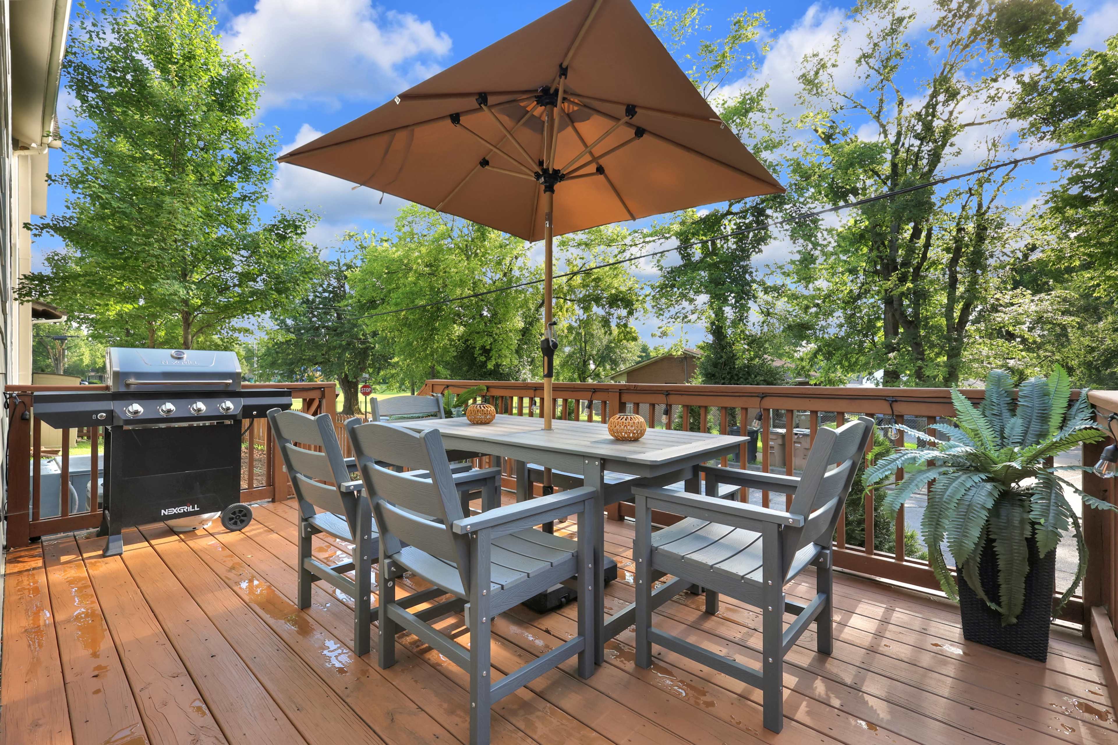 A wooden deck features a dining table with four chairs, an umbrella, and a grill, surrounded by greenery.