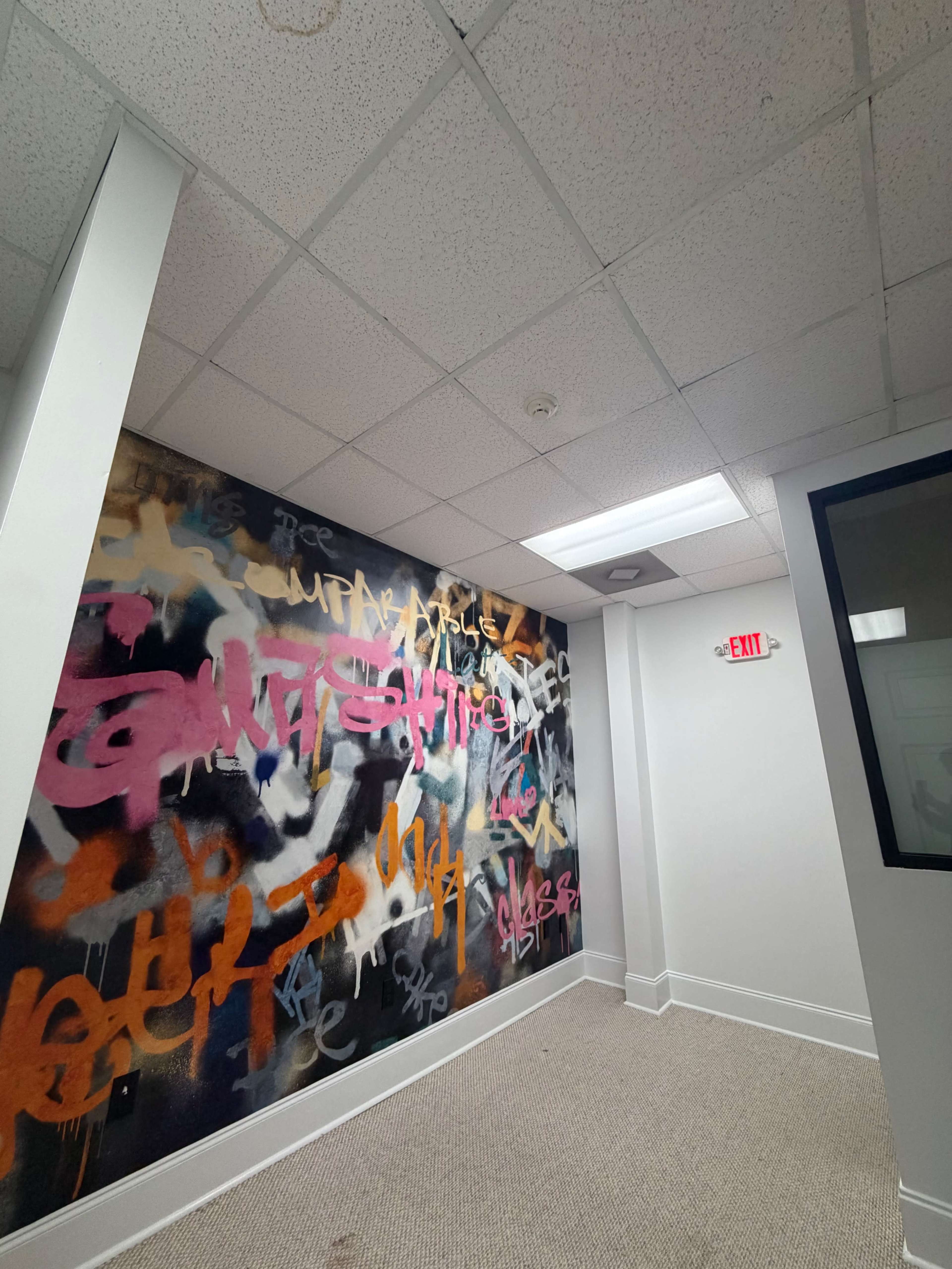 The image shows a corner of a room featuring a wall covered in colorful graffiti with a drop ceiling and an exit sign visible in the background.