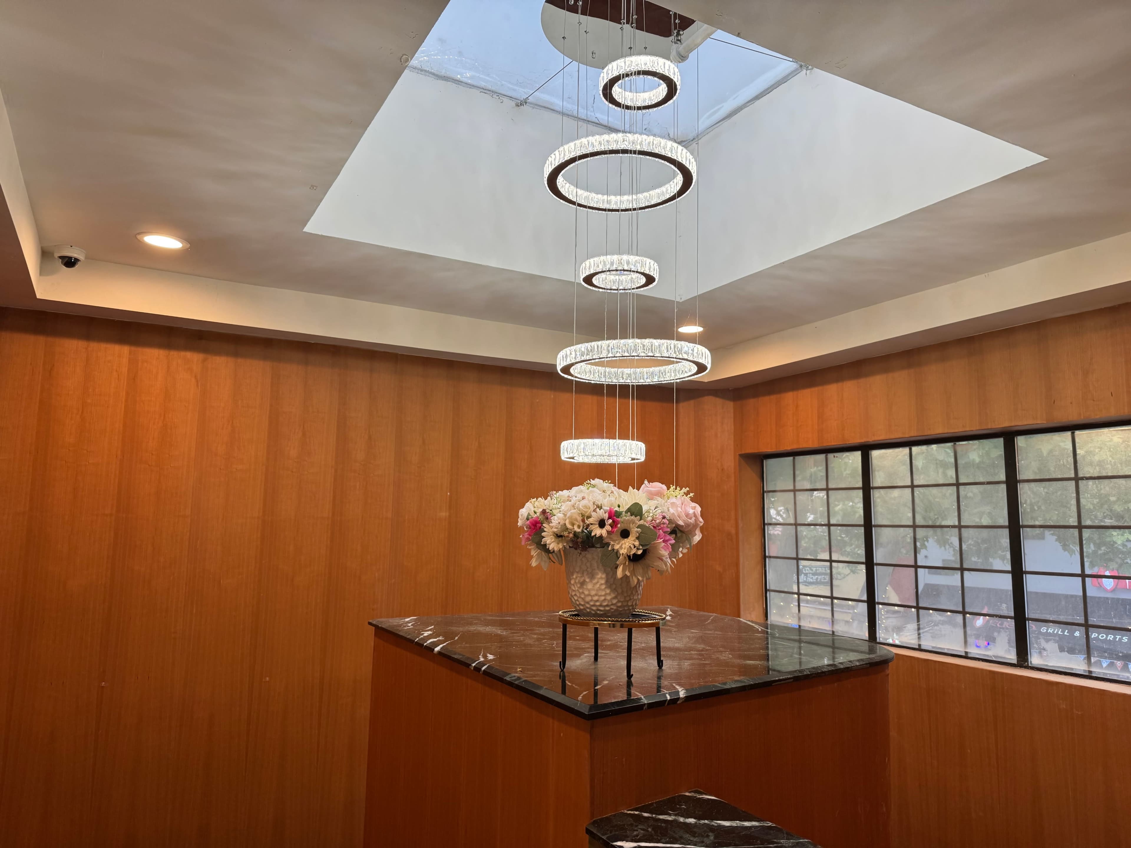 A modern interior features a wooden paneled wall, a central chandelier with circular rings, and a decorative arrangement of flowers on a dark marble counter.
