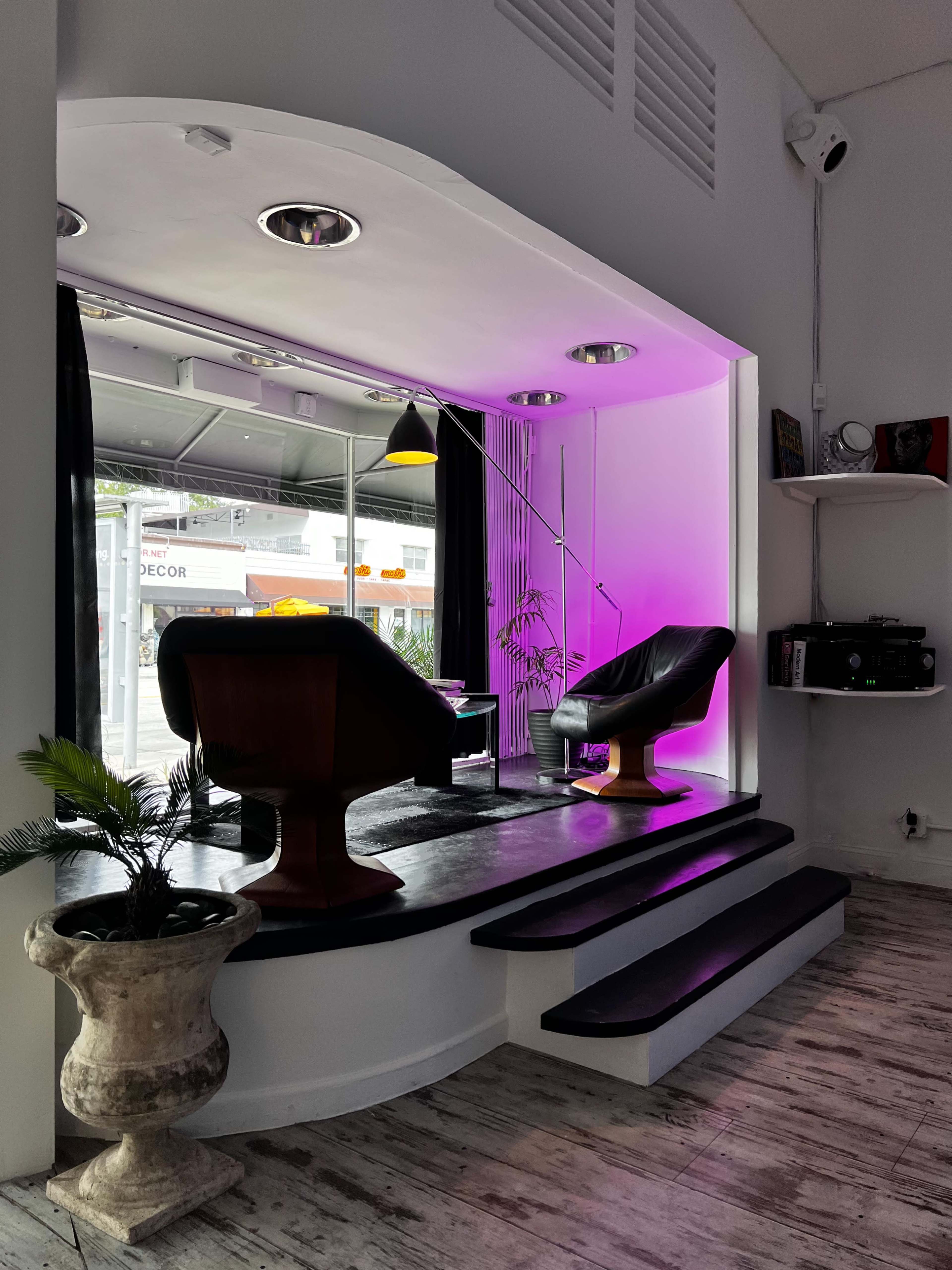 The image shows a modern interior space featuring two chairs on a raised platform with purple lighting, surrounded by large windows and decorative elements.