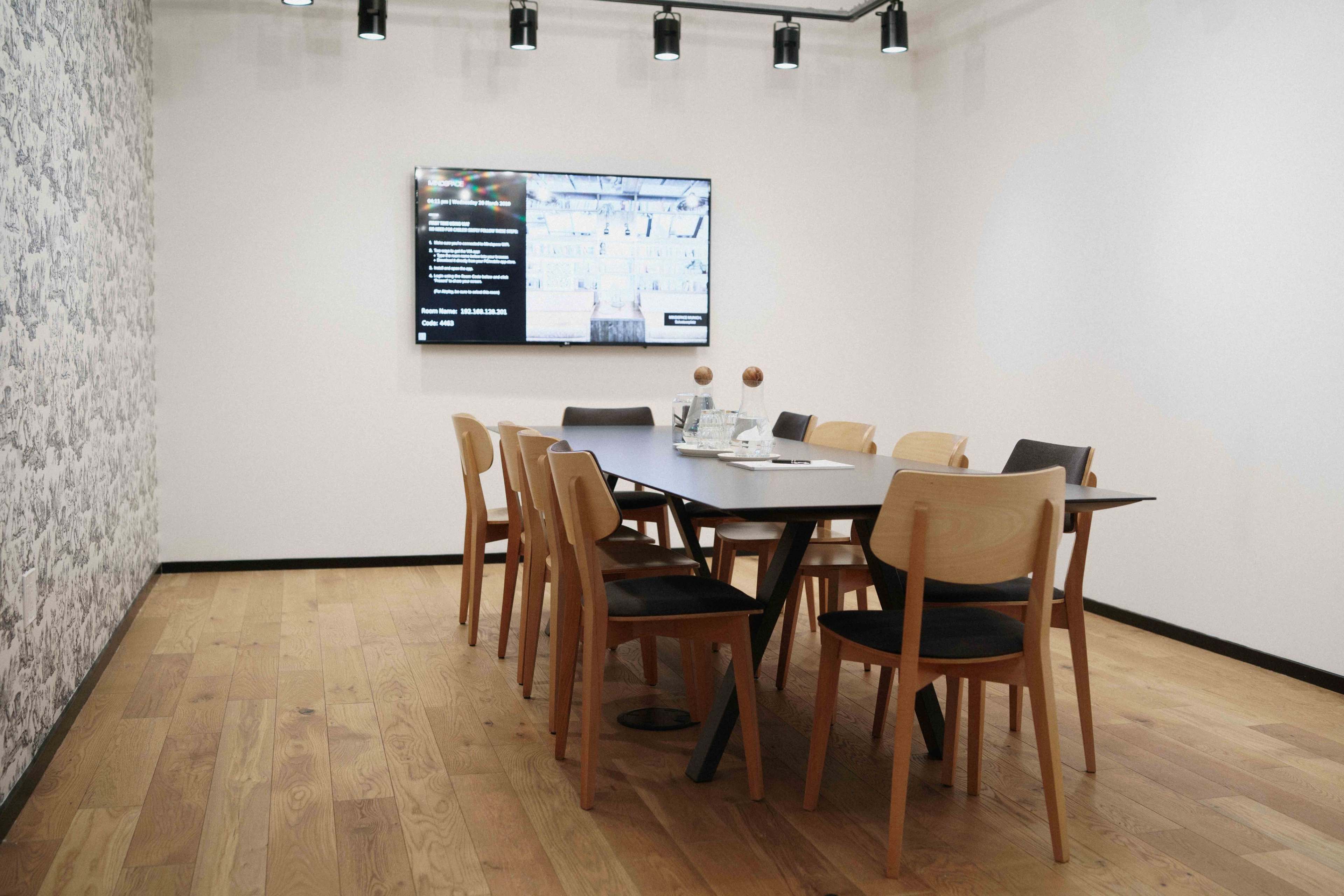 A modern meeting room features a long table surrounded by wooden chairs and a large screen displaying information on a white wall.