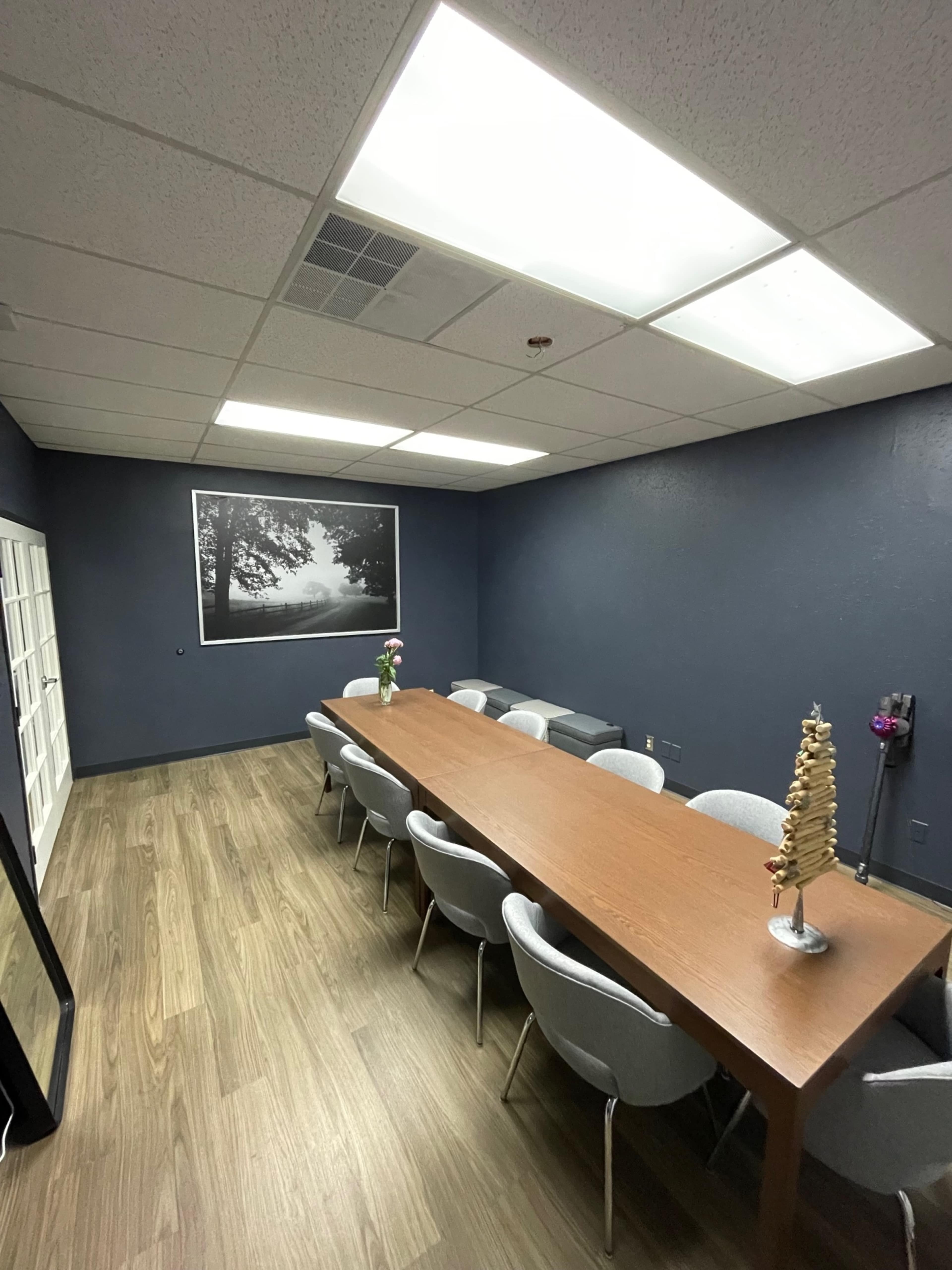 A conference room features a long wooden table surrounded by gray chairs, with a large framed black-and-white photo on the wall.
