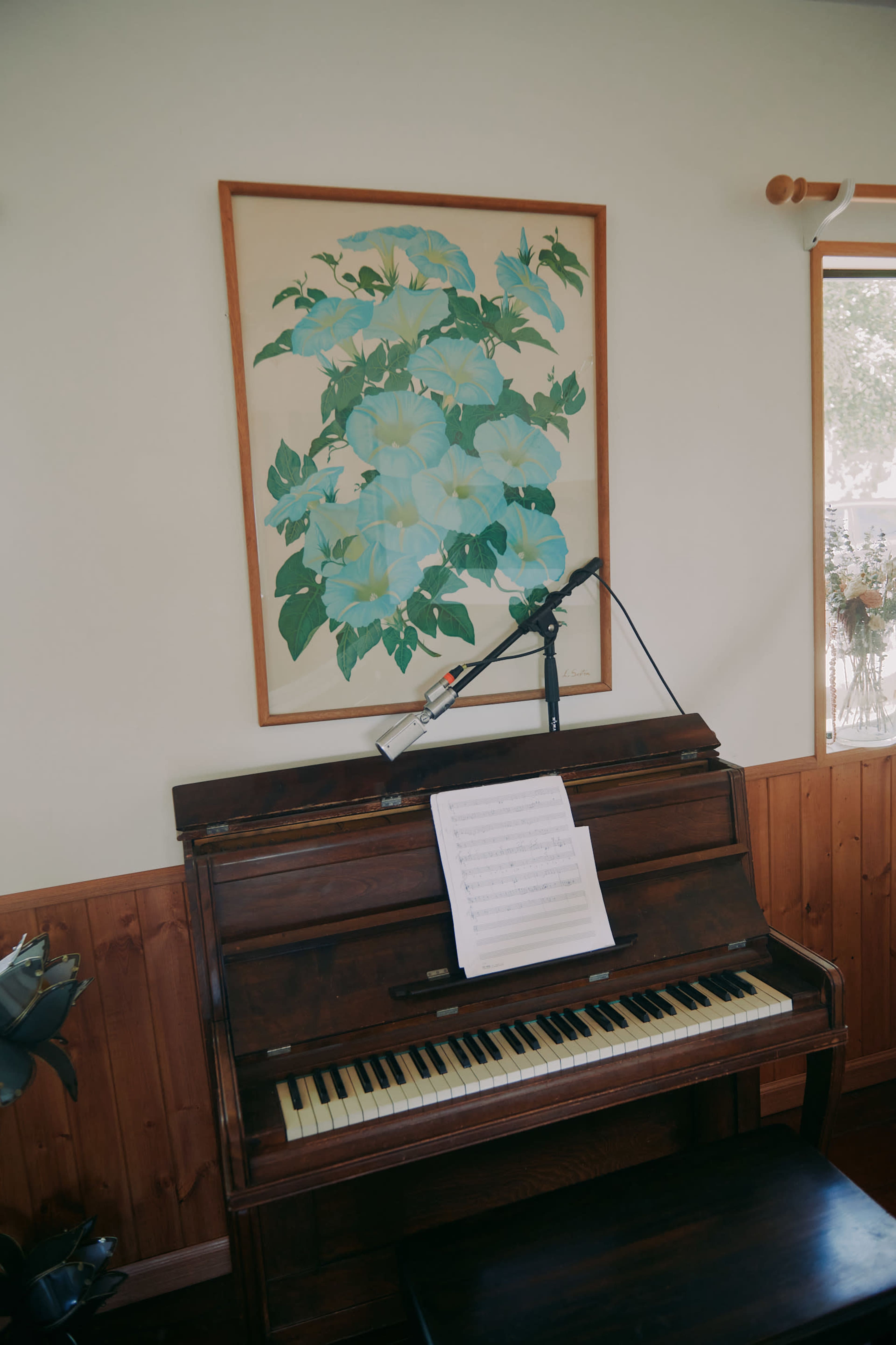 A vintage piano sits against a wooden wall, with a large floral painting above it and a microphone positioned nearby.