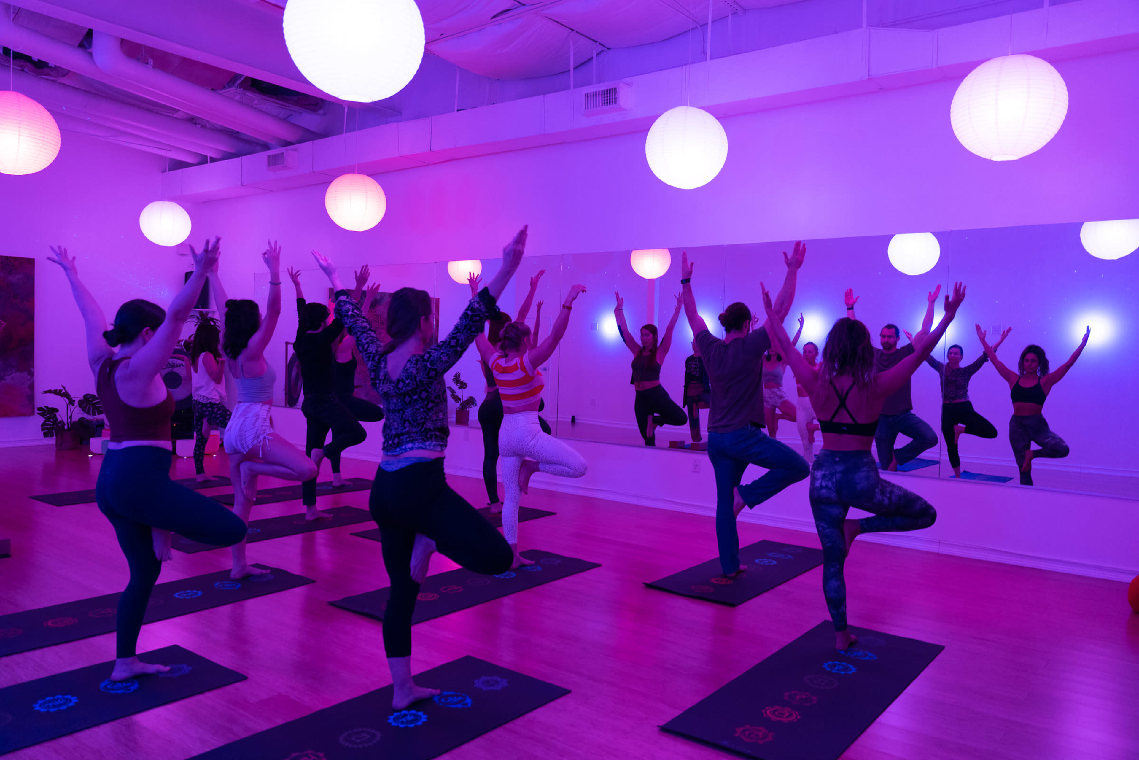A group of people practices yoga in a brightly lit studio with purple and white hanging lamps.