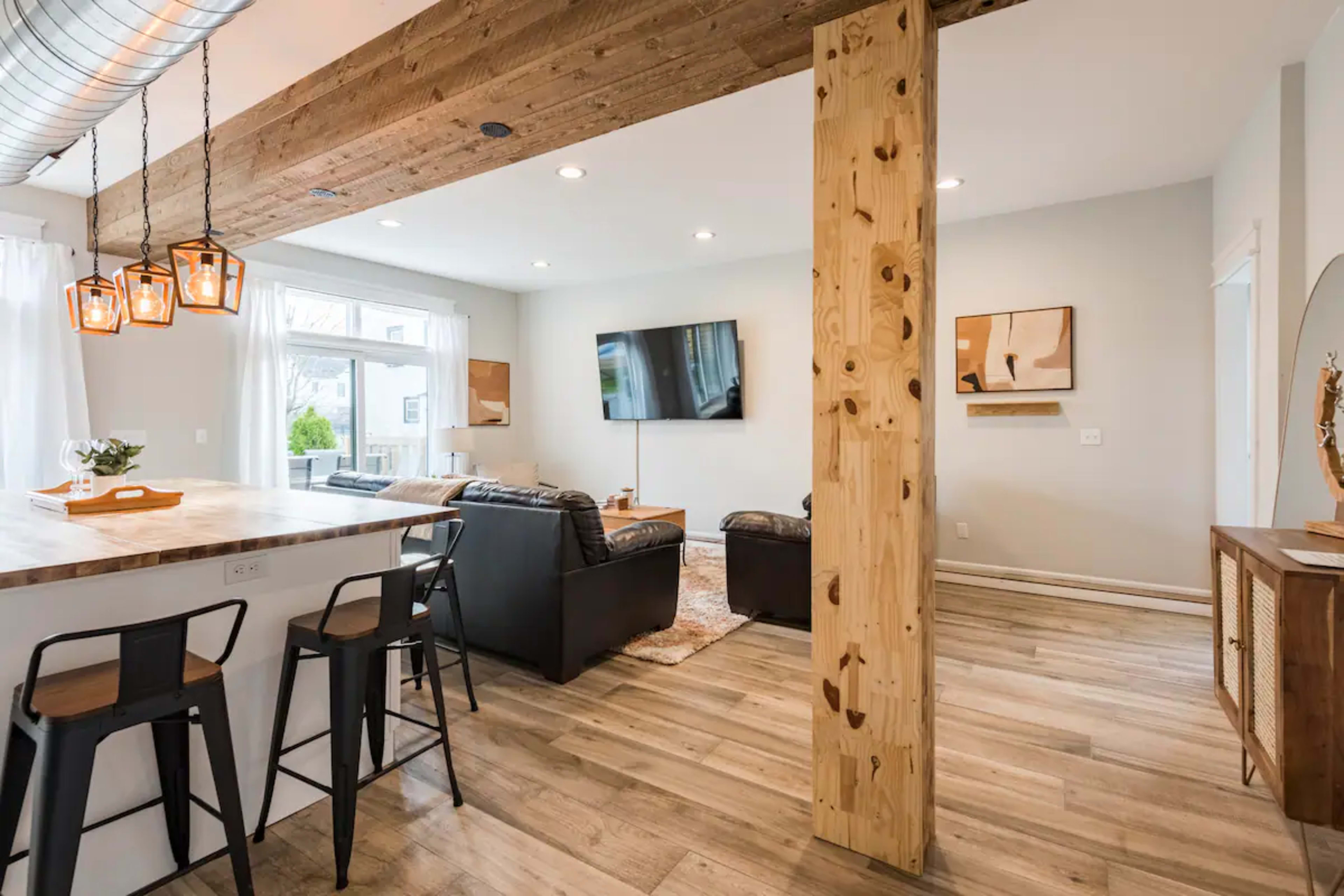 The image shows a modern living space featuring a dining area with bar stools, a cozy seating arrangement, a television mounted on the wall, and wooden accents throughout the room.