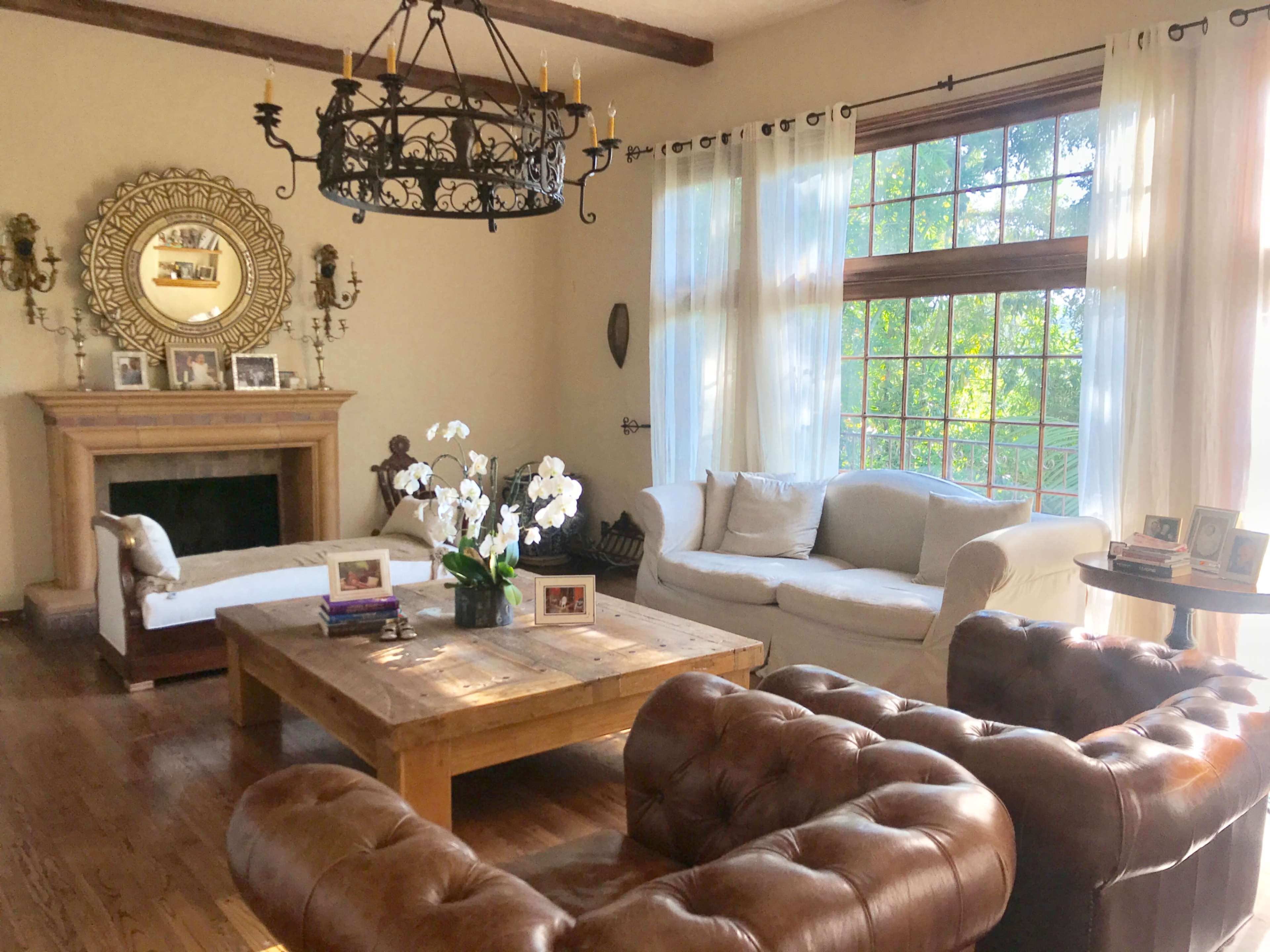 A cozy living room featuring a wooden coffee table, a white couch, a brown leather chair, and large windows with sheer curtains.