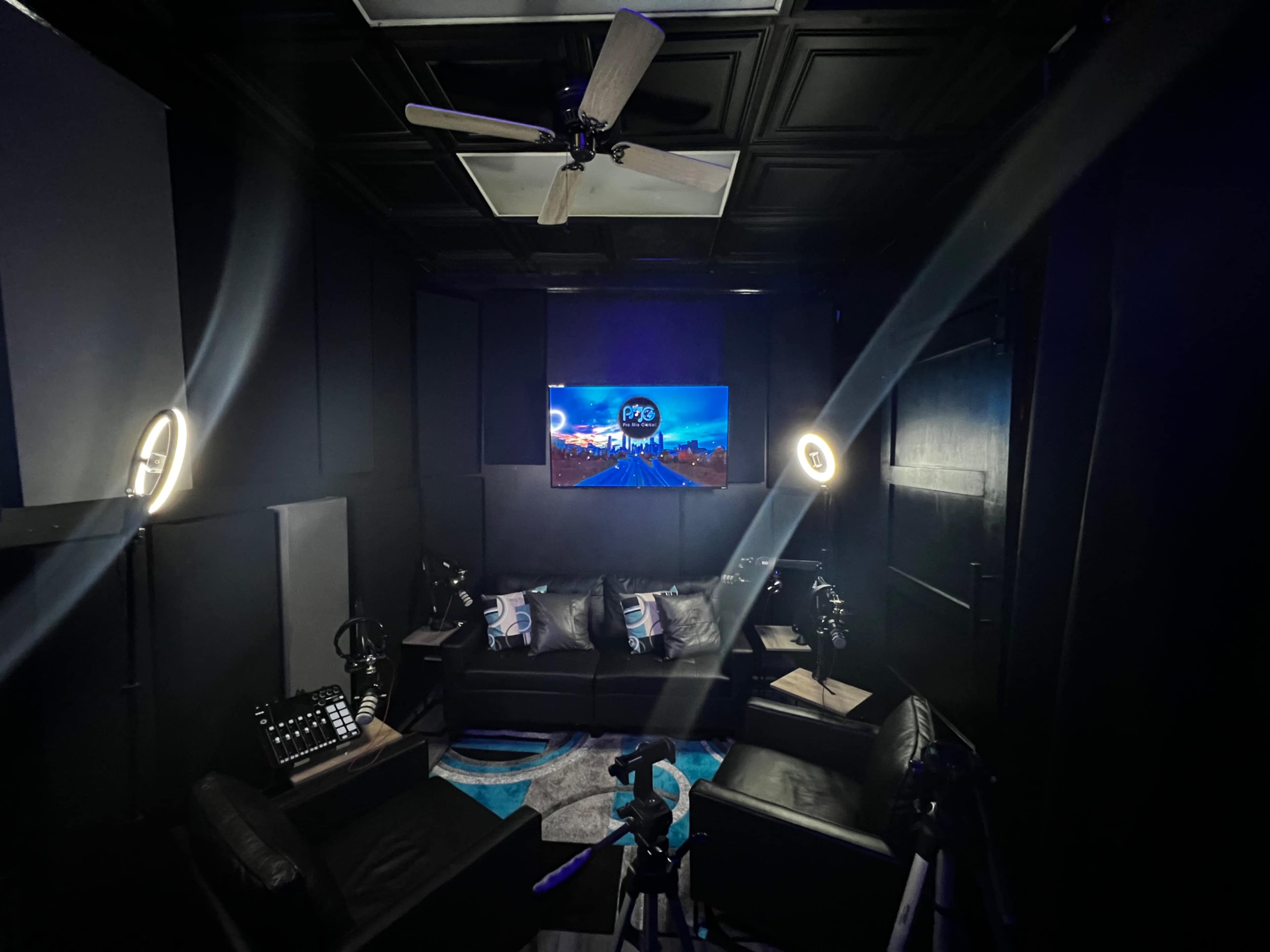 A dark studio with black walls, a large TV displaying a cityscape, and professional lighting setups surrounding a seating area.