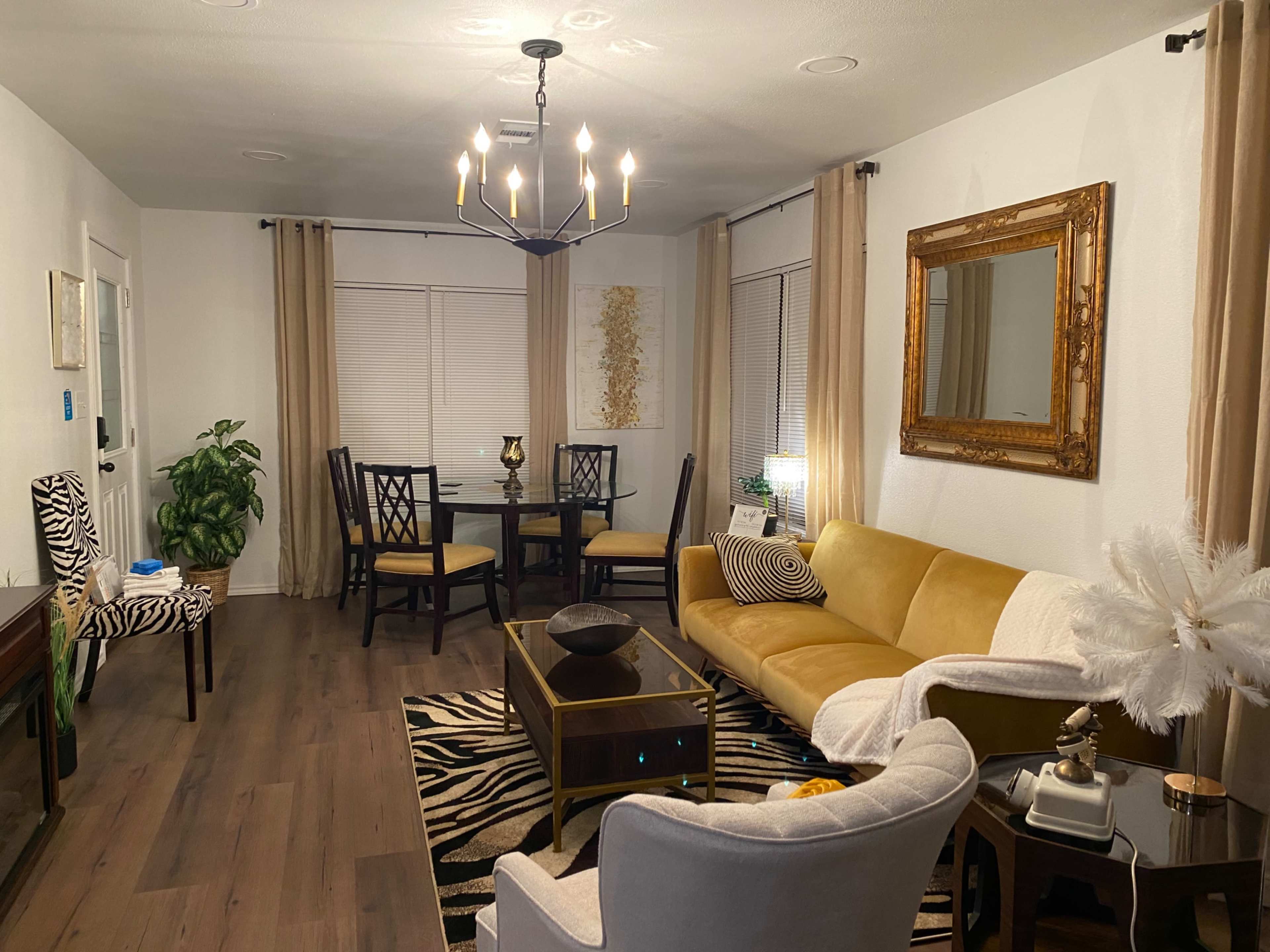 A modern living area with a yellow couch, a glass coffee table, a dining table in the background, and decor featuring animal patterns and plants.