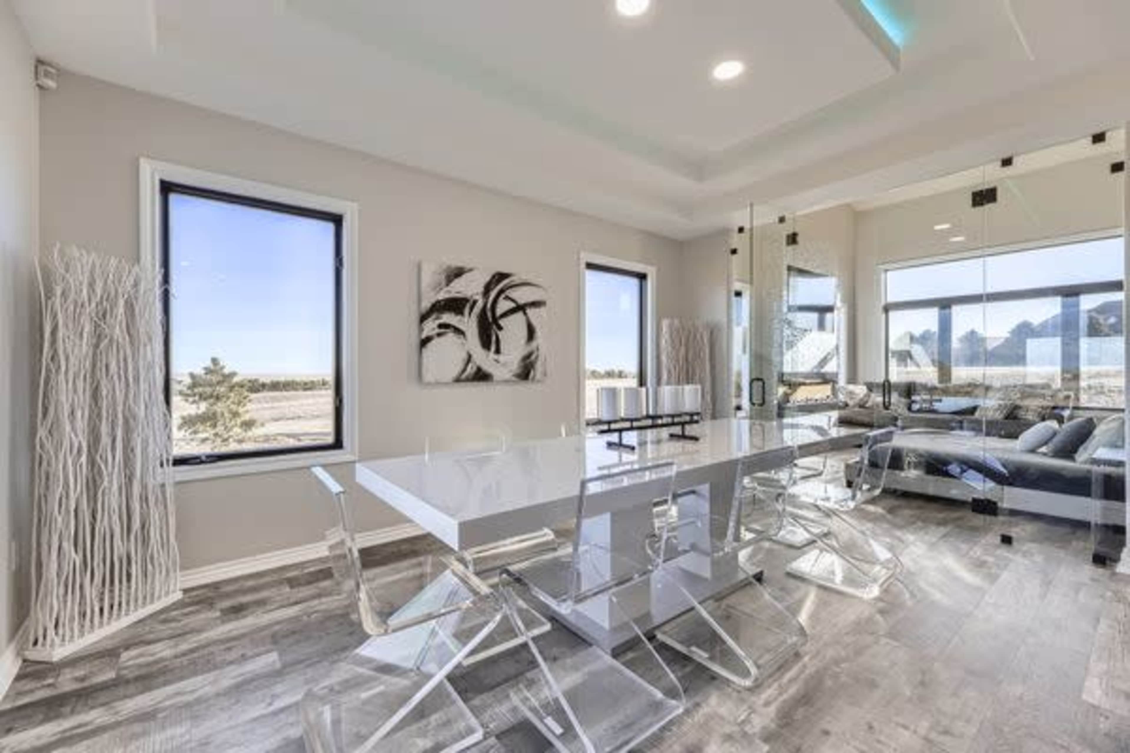 The image shows a modern dining area with a sleek white table surrounded by transparent chairs, large windows revealing an outdoor view, and minimalist decor.
