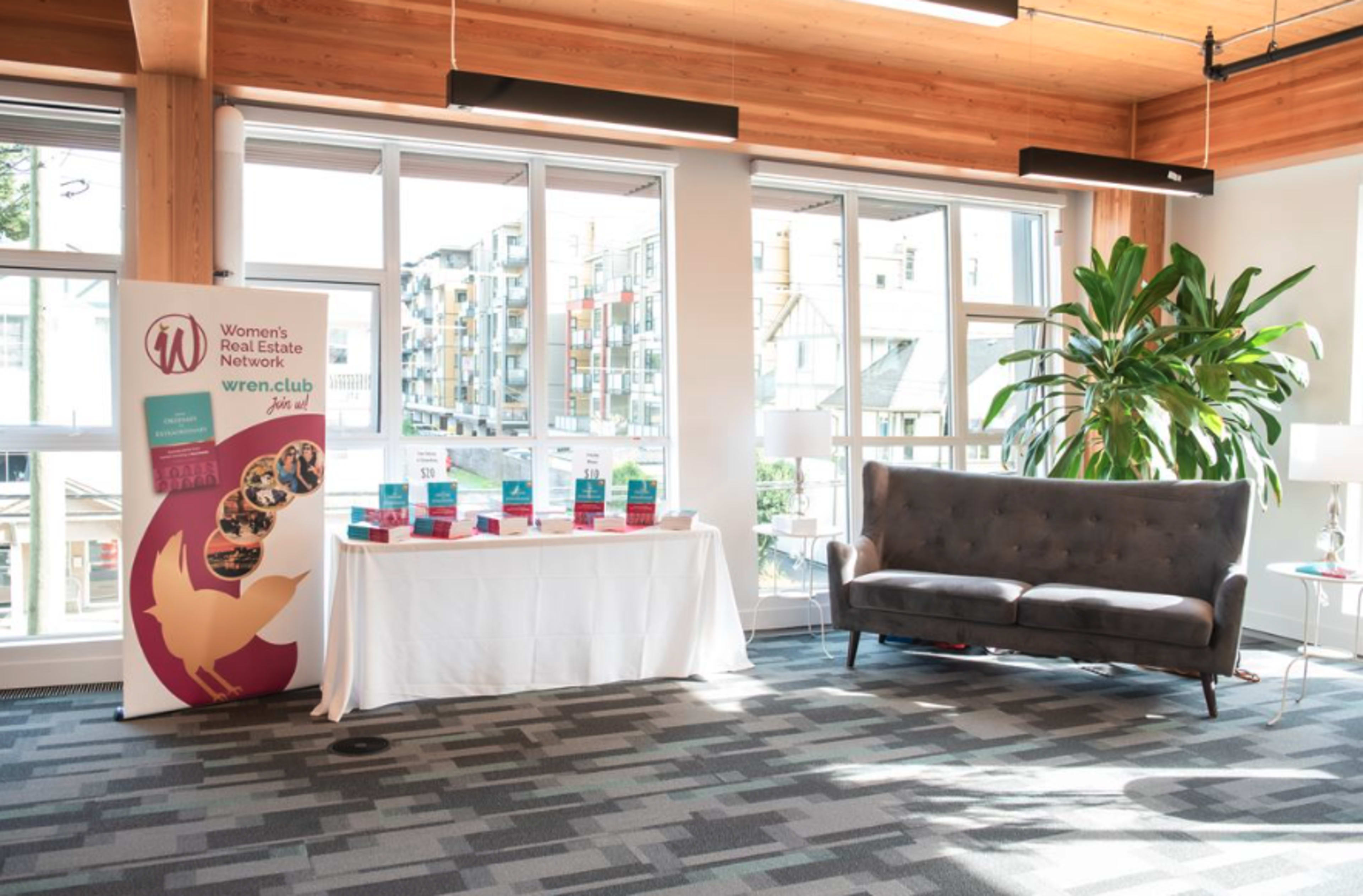 A furnished interior space featuring a banner for the Women's Real Estate Network, a table with promotional materials, and a couch near large windows.