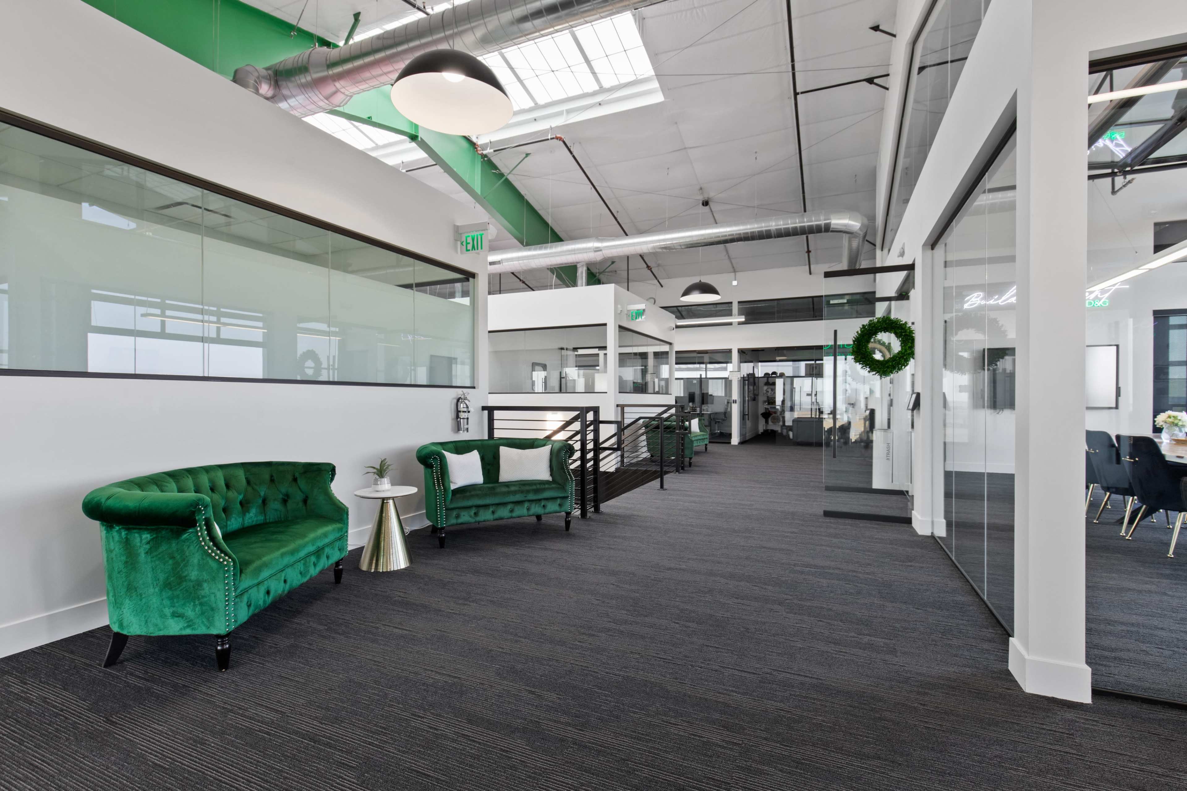 The image shows a modern office corridor featuring green velvet couches, glass-walled meeting rooms, and sleek industrial design elements.