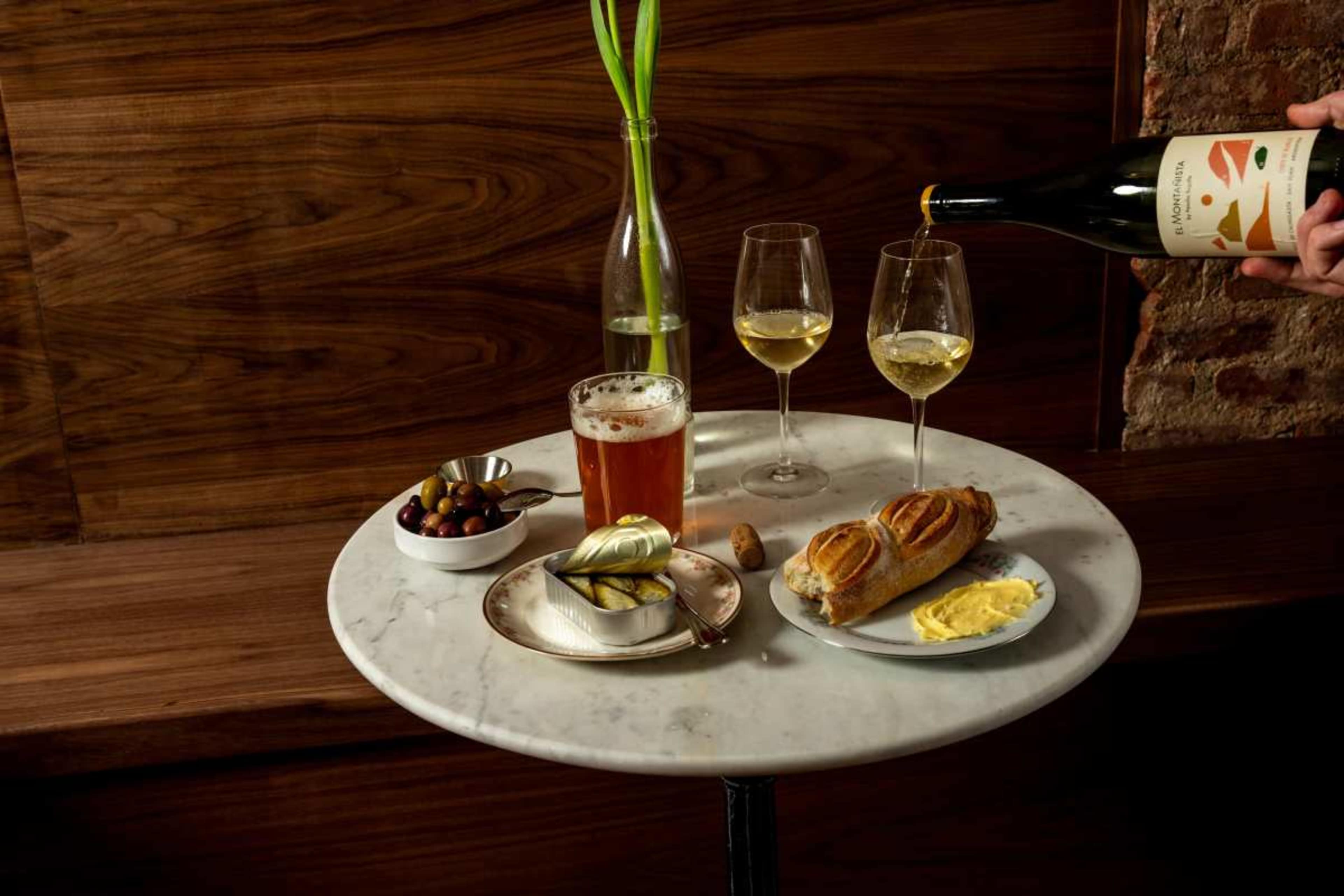 A marble table holds two glasses of white wine, a glass of beer, a plate of butter and bread, a dish of olives, and a can of sardines, alongside a tall vase with green plants.