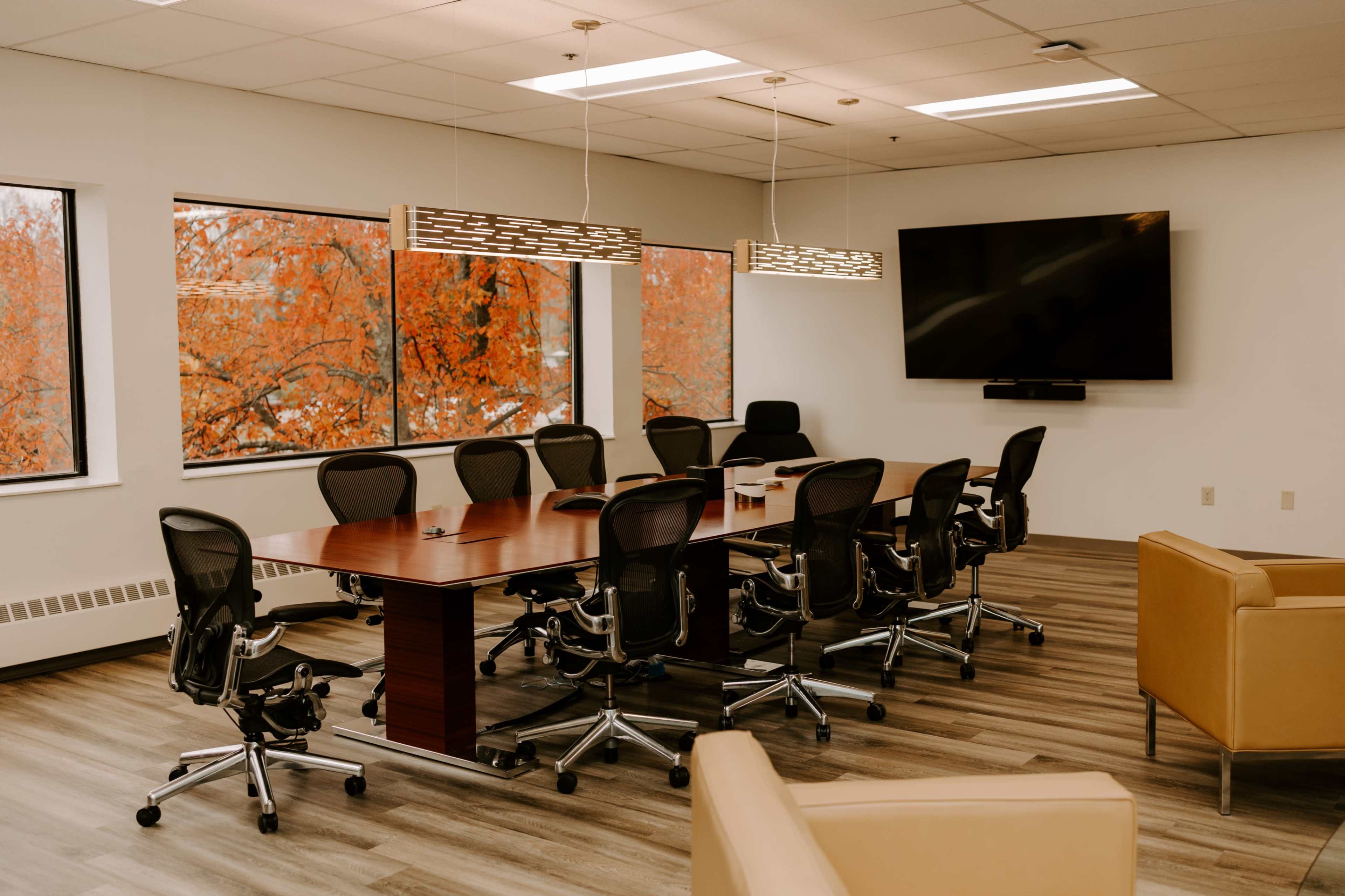 A conference room features a large wooden table surrounded by ergonomic chairs, with large windows displaying fall foliage outside.
