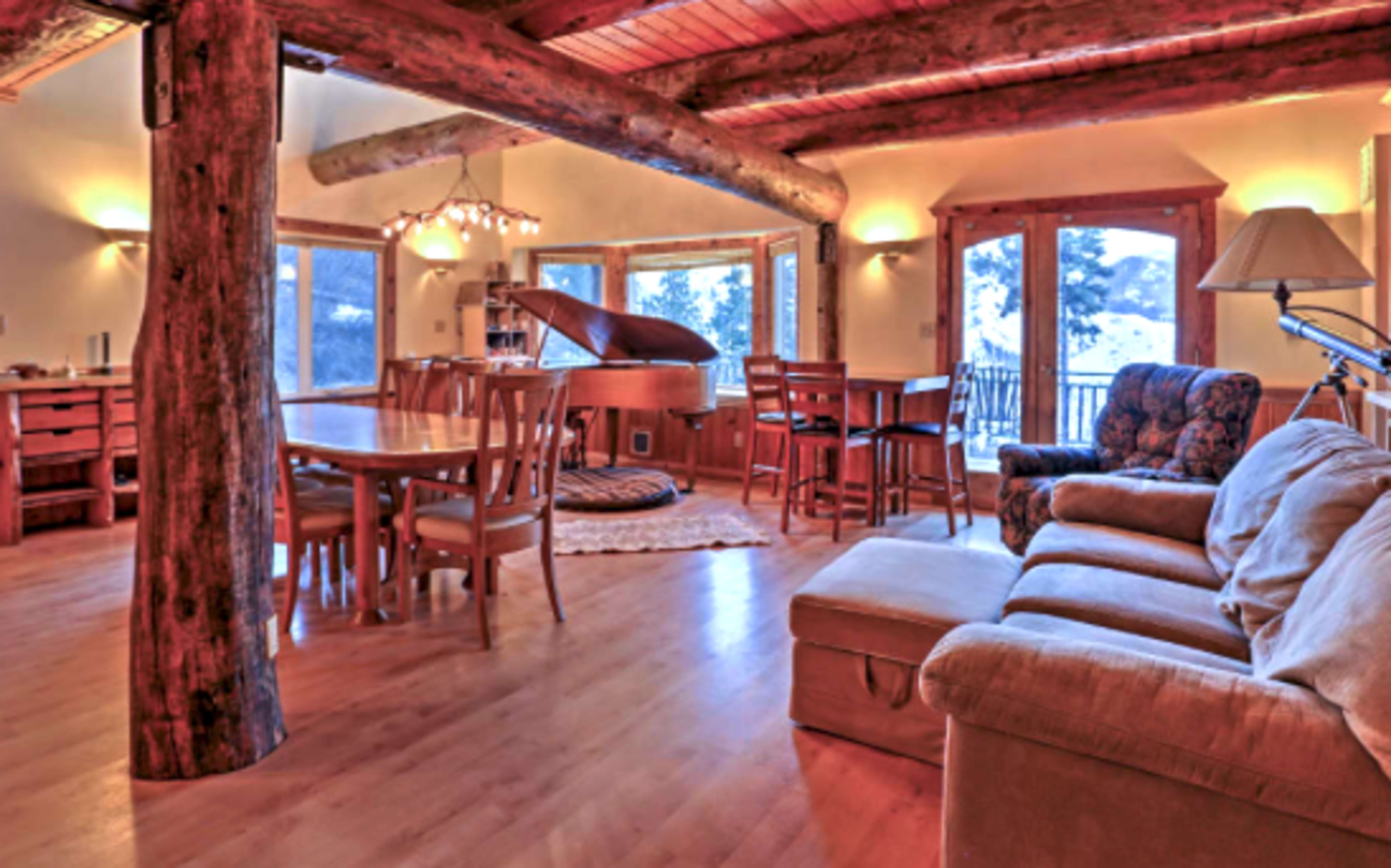 A spacious living area featuring a grand piano, a dining table, and large windows that offer an outdoor view, all set against wooden beams and a warm interior design.