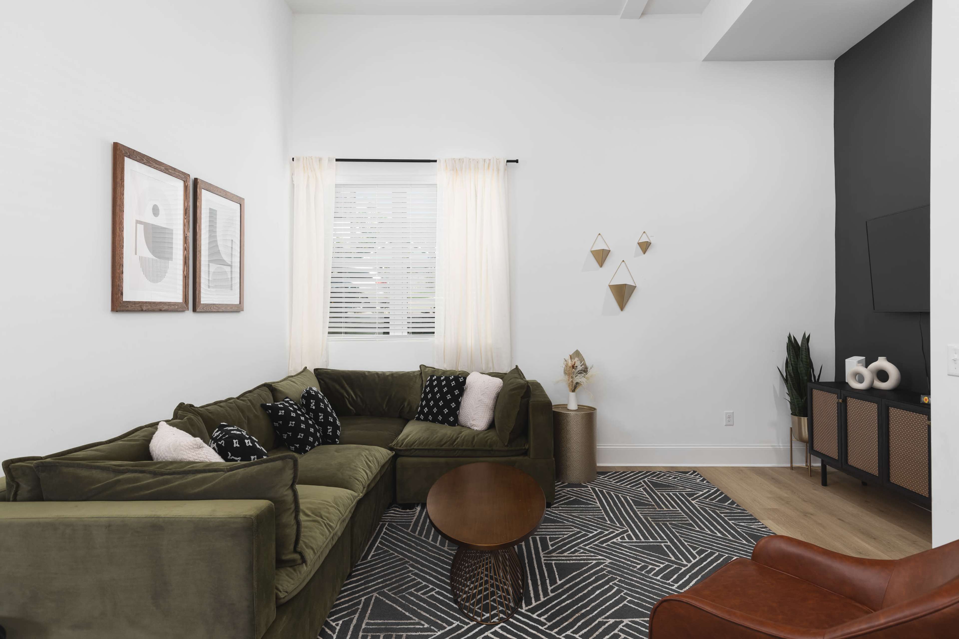 A modern living room featuring a green sectional sofa, a round wooden coffee table, framed artwork on the wall, and geometric decor.