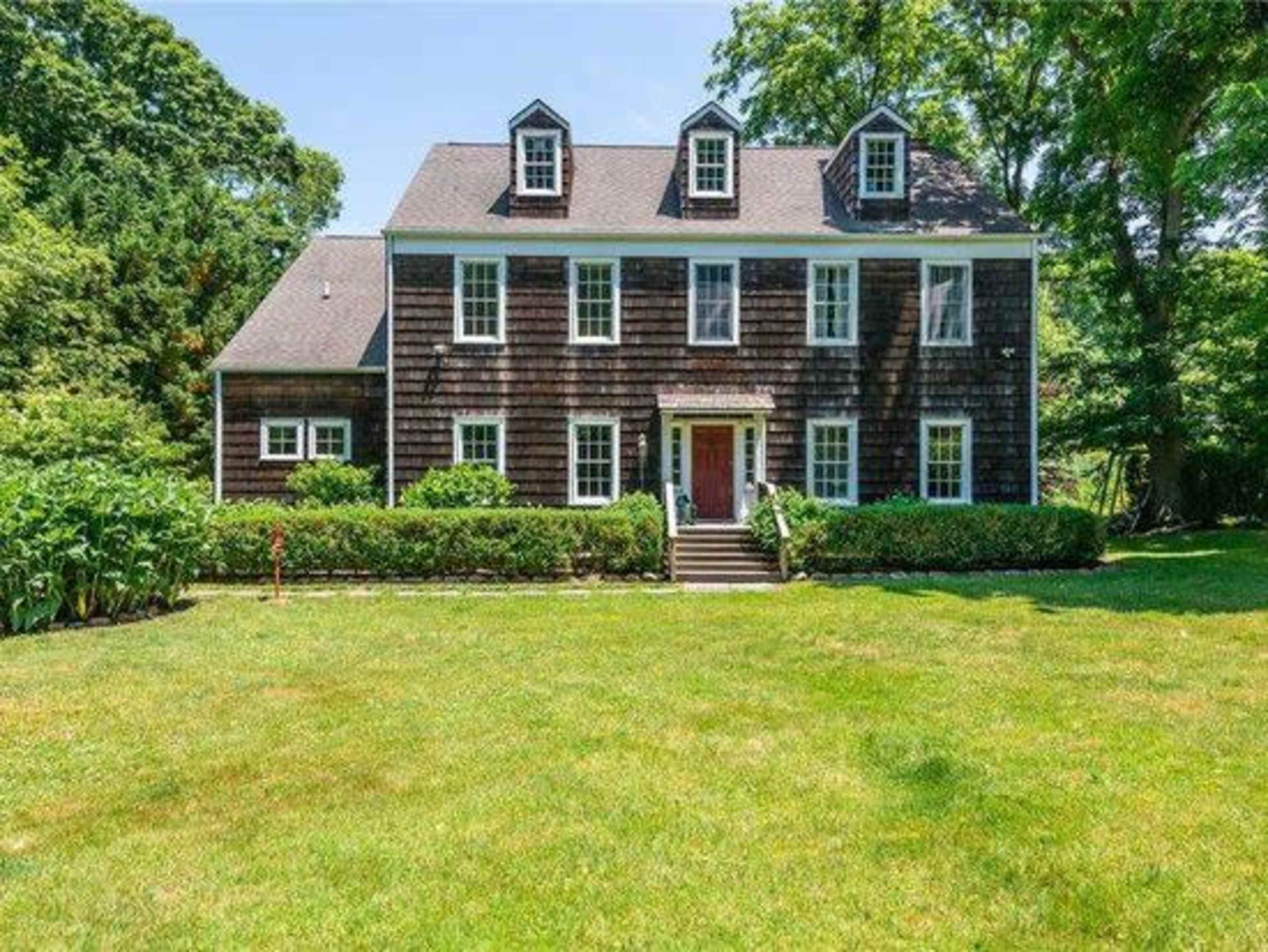 A two-story wooden house with a front porch, surrounded by a well-maintained lawn and dense greenery.