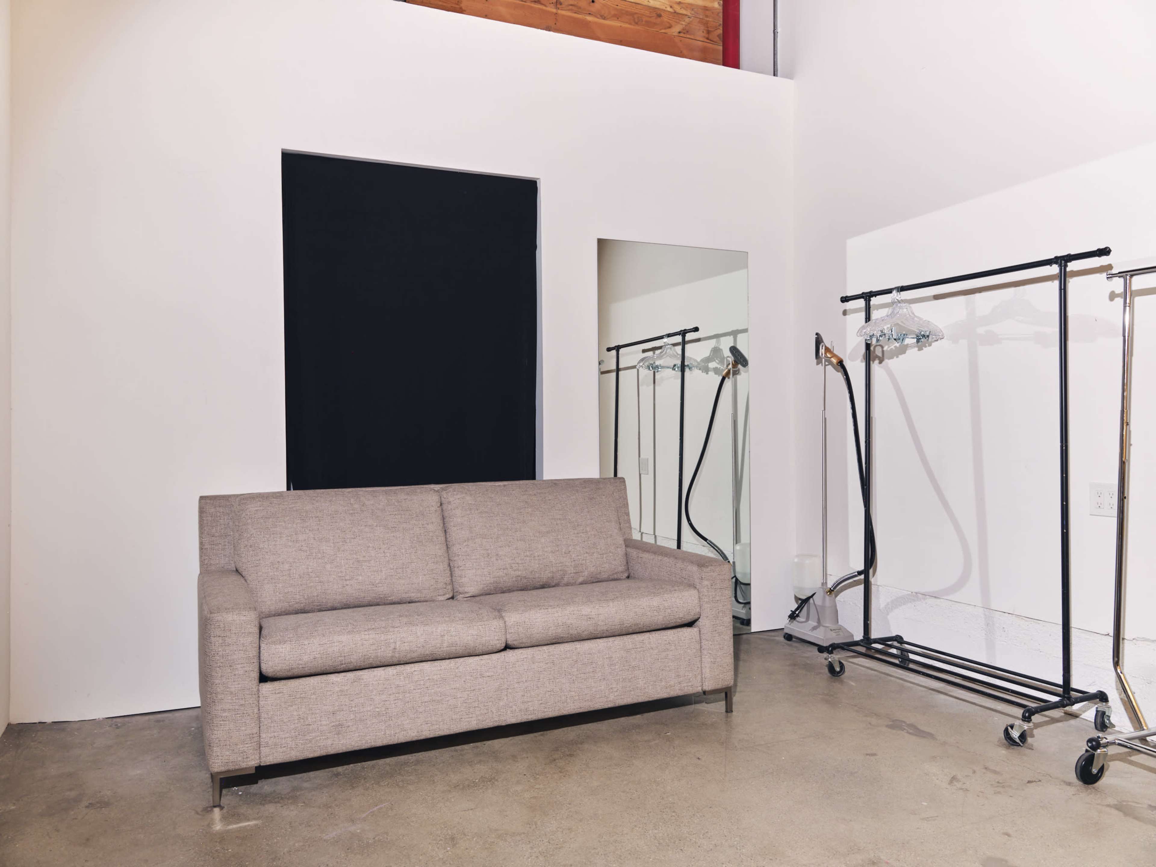 The image shows a beige sofa situated against a white wall with a black window and a mirror, alongside clothing racks on wheels.