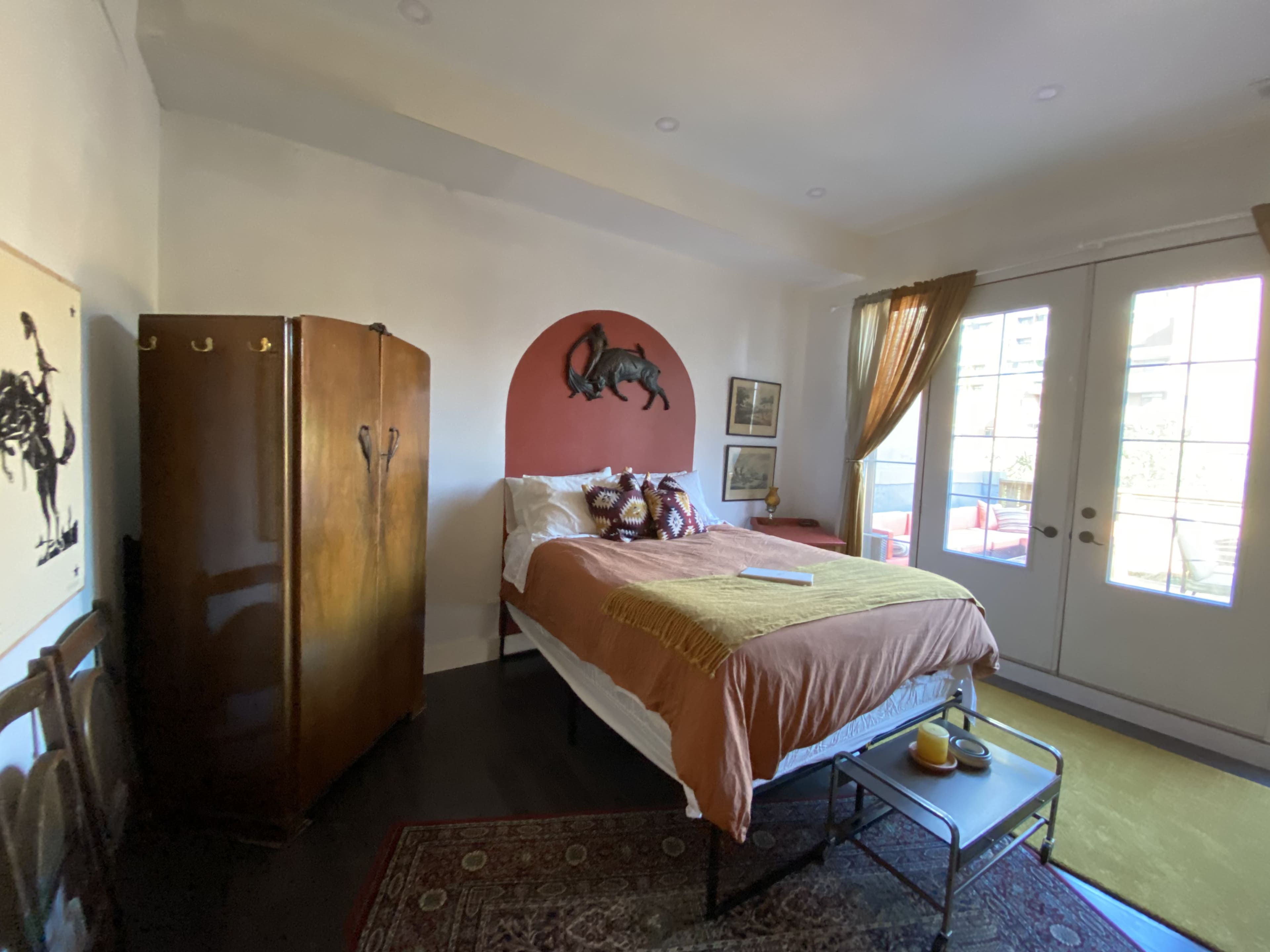 A bedroom features a bed with orange bedding, a wooden wardrobe, and a decorative wall with a horse painting, along with large glass doors opening to an outdoor area.