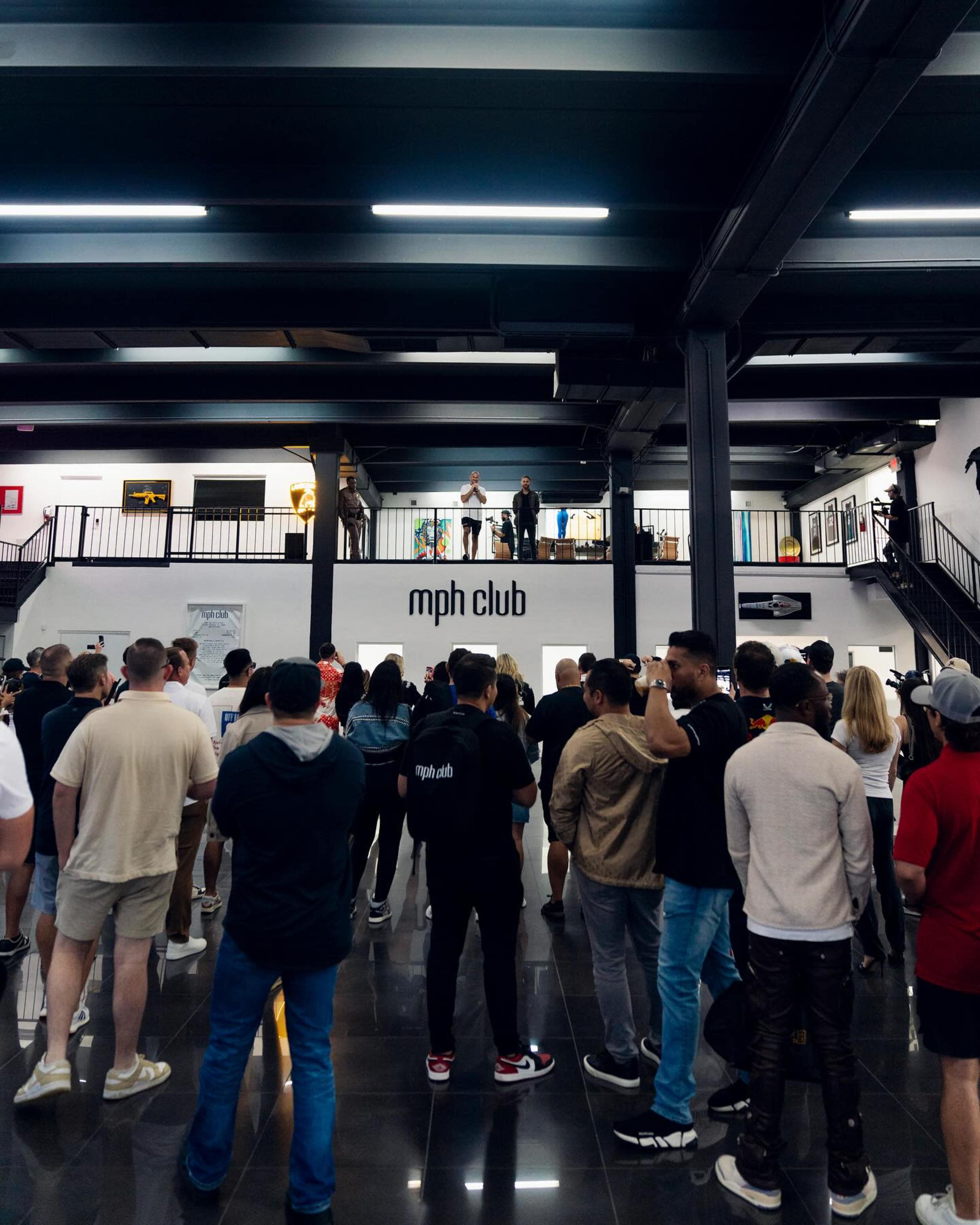 A crowd gathers in a large indoor space under black beams, listening to a speaker on an upper level with a sign for "mph club" in the background.