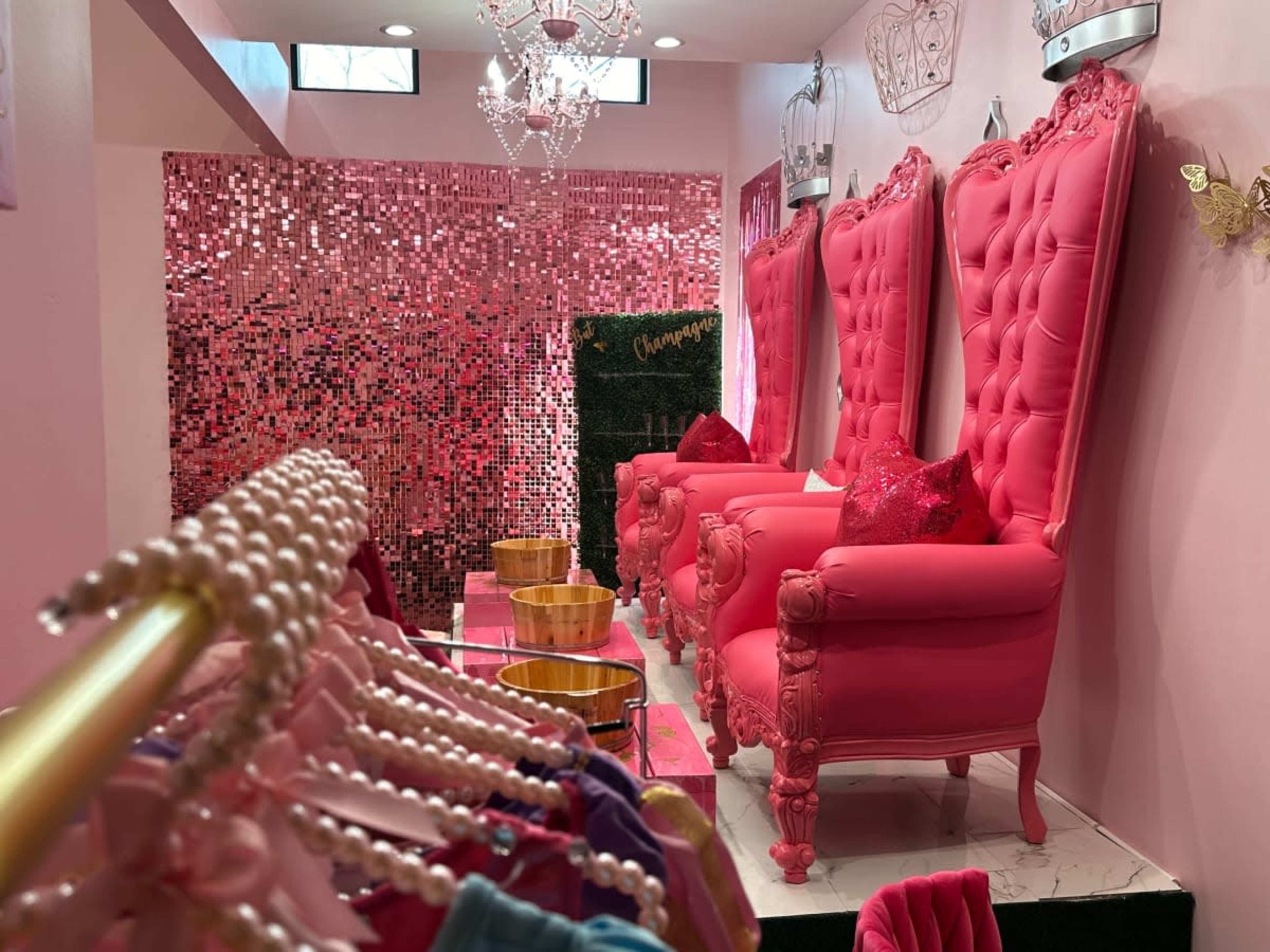 A stylish boutique interior featuring three large pink chairs, a shimmering pink wall, and a display of colorful clothing on hangers.