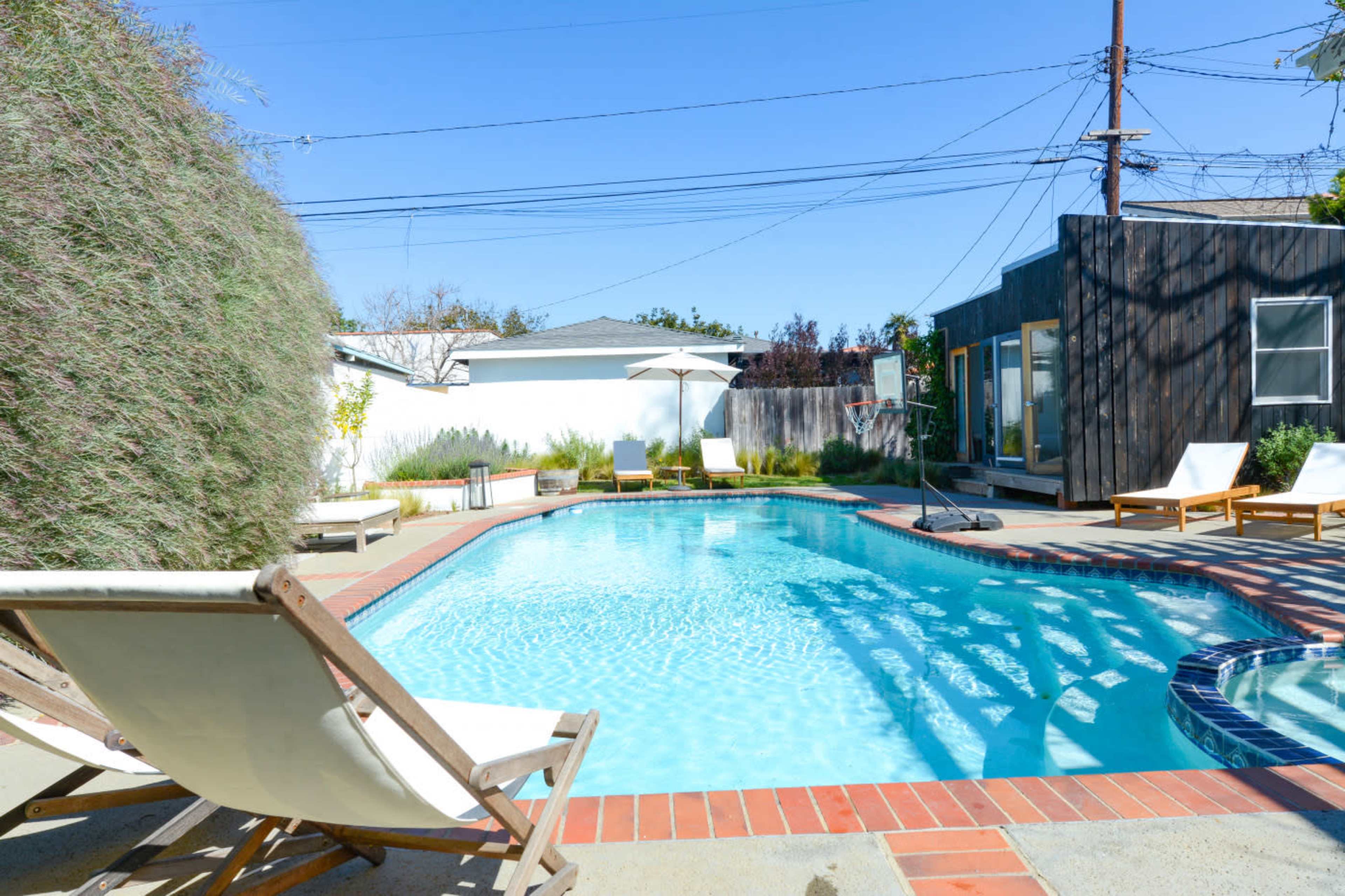 A blue swimming pool with lounge chairs nearby, surrounded by a landscaped area and a wooden structure in the background.
