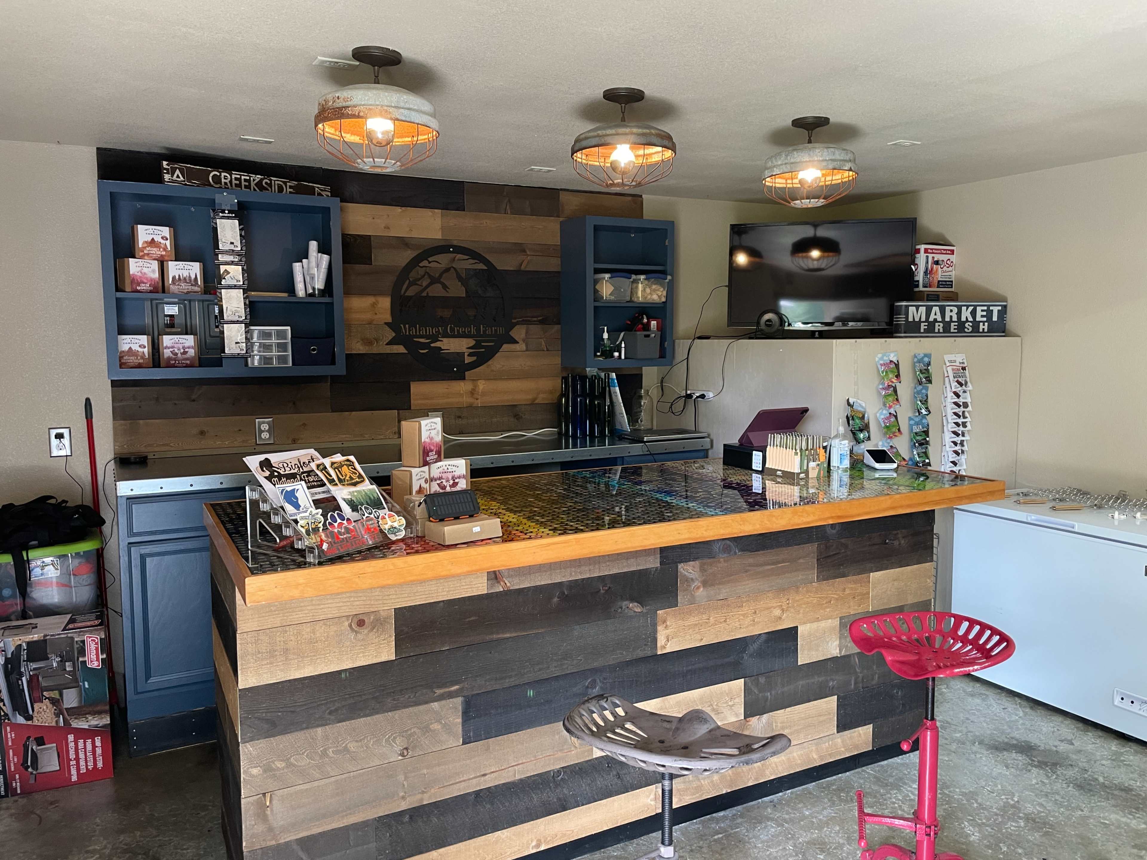 A small, rustic bar area with a wooden counter, shelves displaying various items, and a television mounted on the wall.