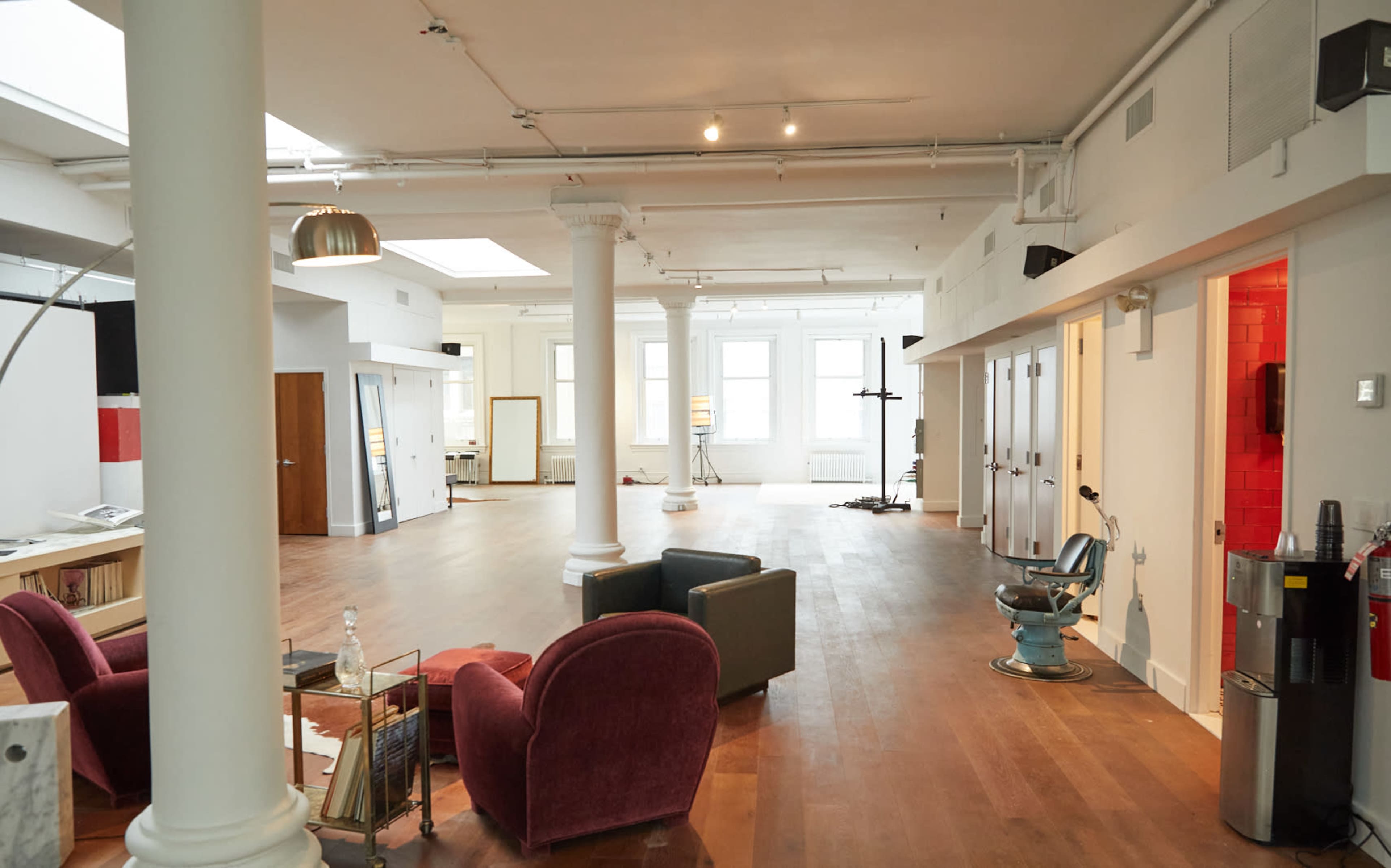 The image shows a spacious, well-lit open-plan interior featuring hardwood floors, several columns, and a mix of seating arrangements including two armchairs and a small table.