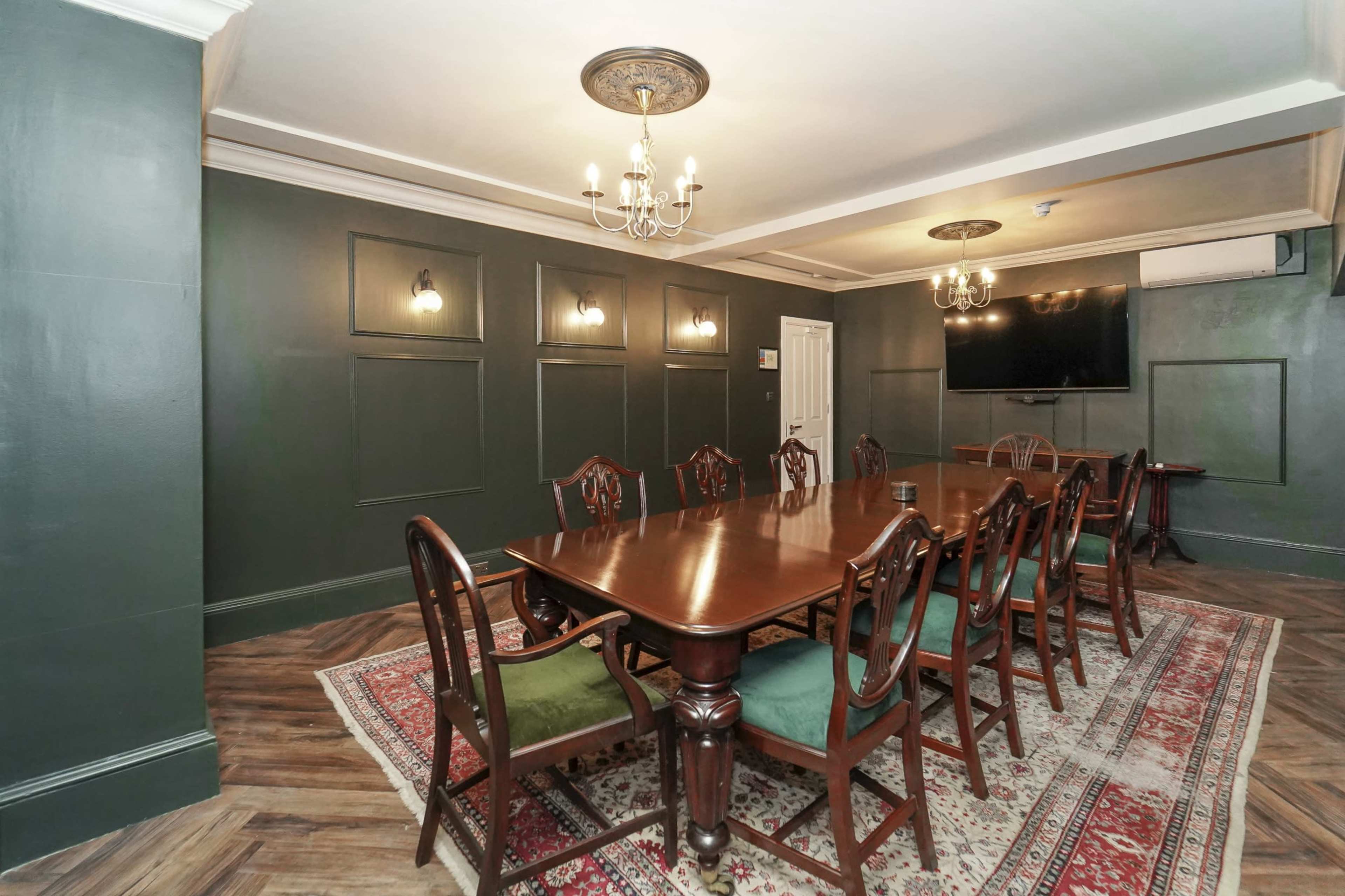 The image shows a dining room with a long wooden table surrounded by chairs, set against dark green walls and featuring a decorative chandelier and wall-mounted television.