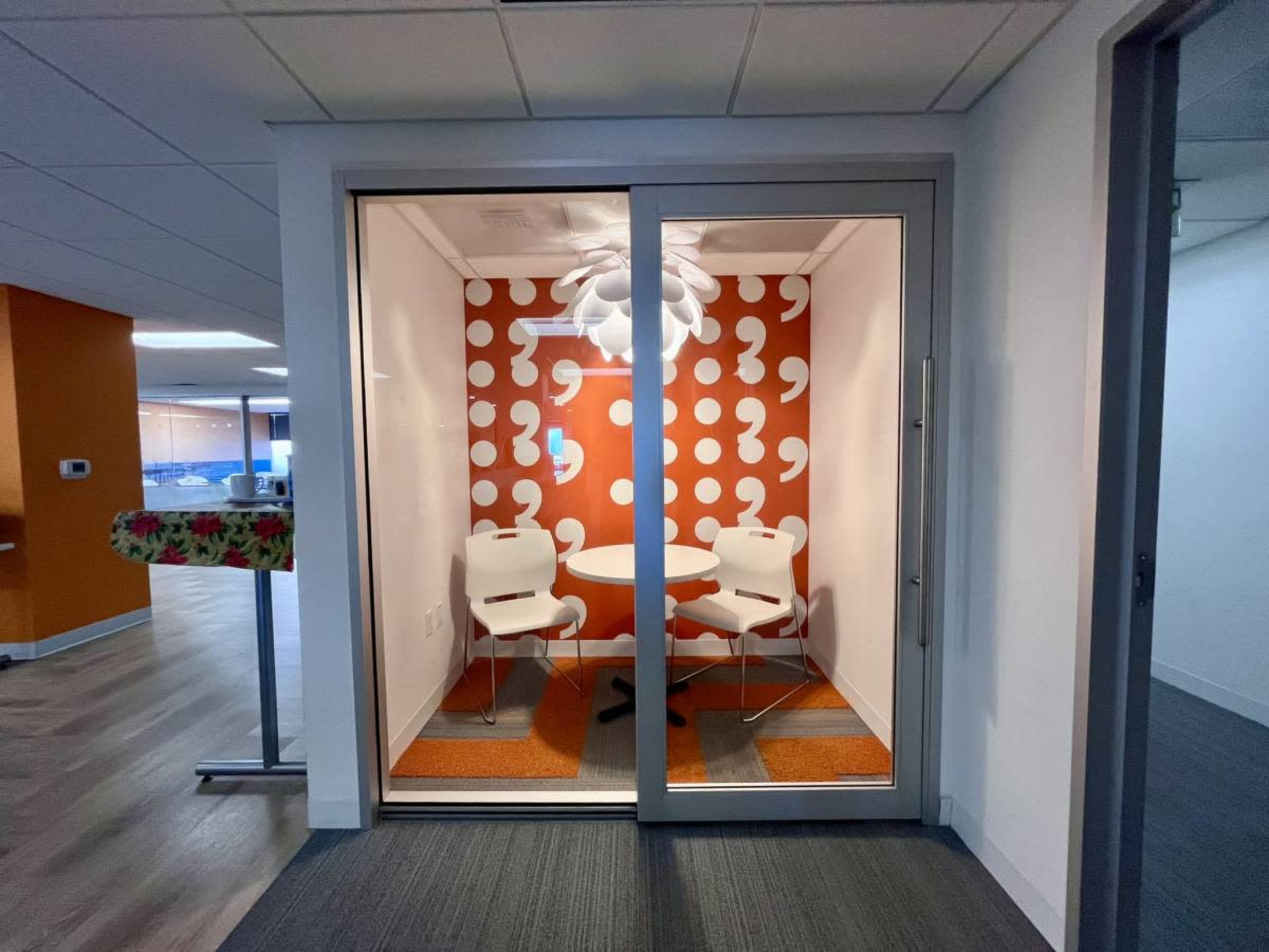 A small office meeting room with a round table and two chairs, featuring an orange wall with white circular patterns.