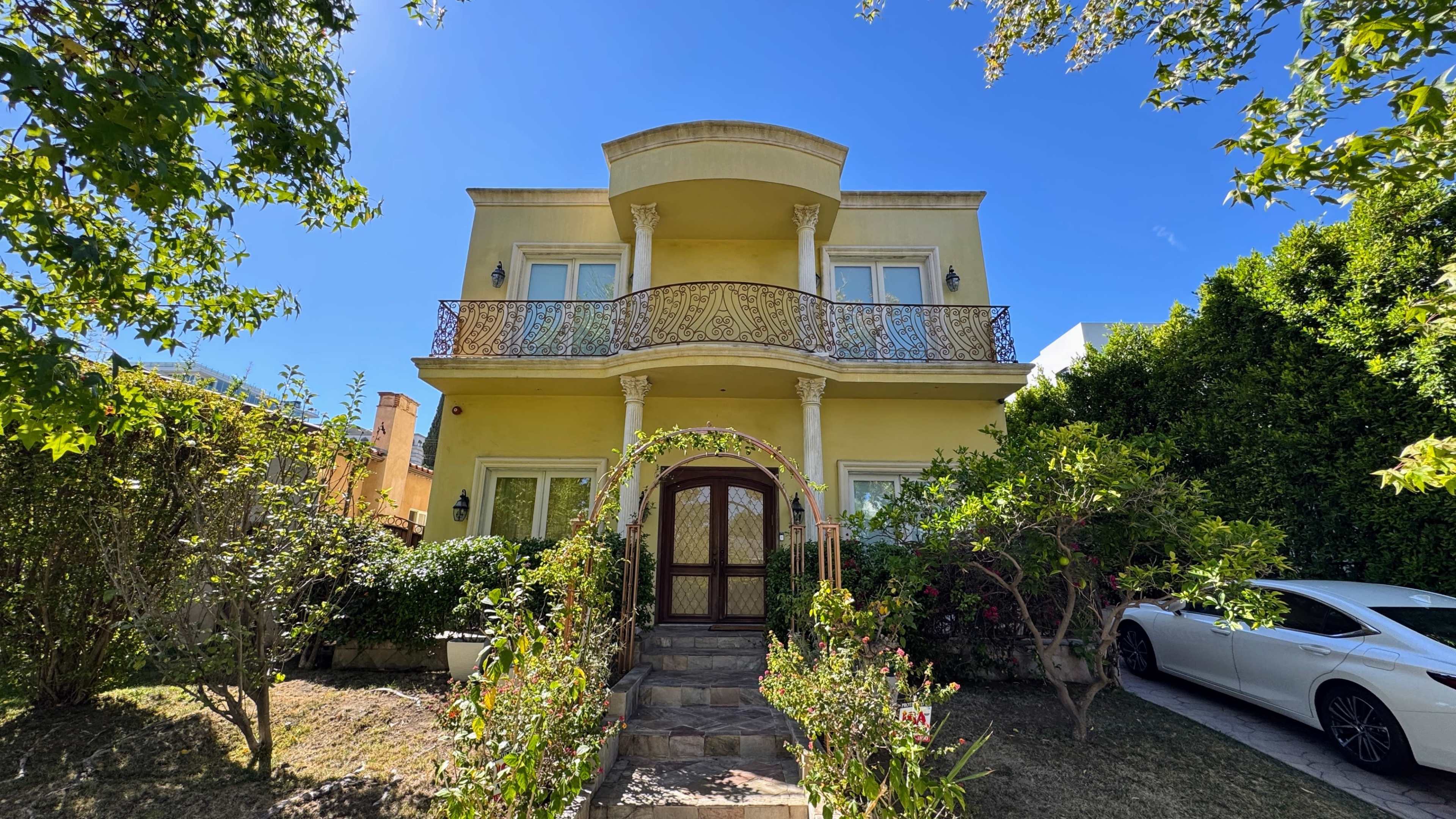 A two-story yellow house with ornate balconies and a landscaped garden is situated under a clear blue sky.