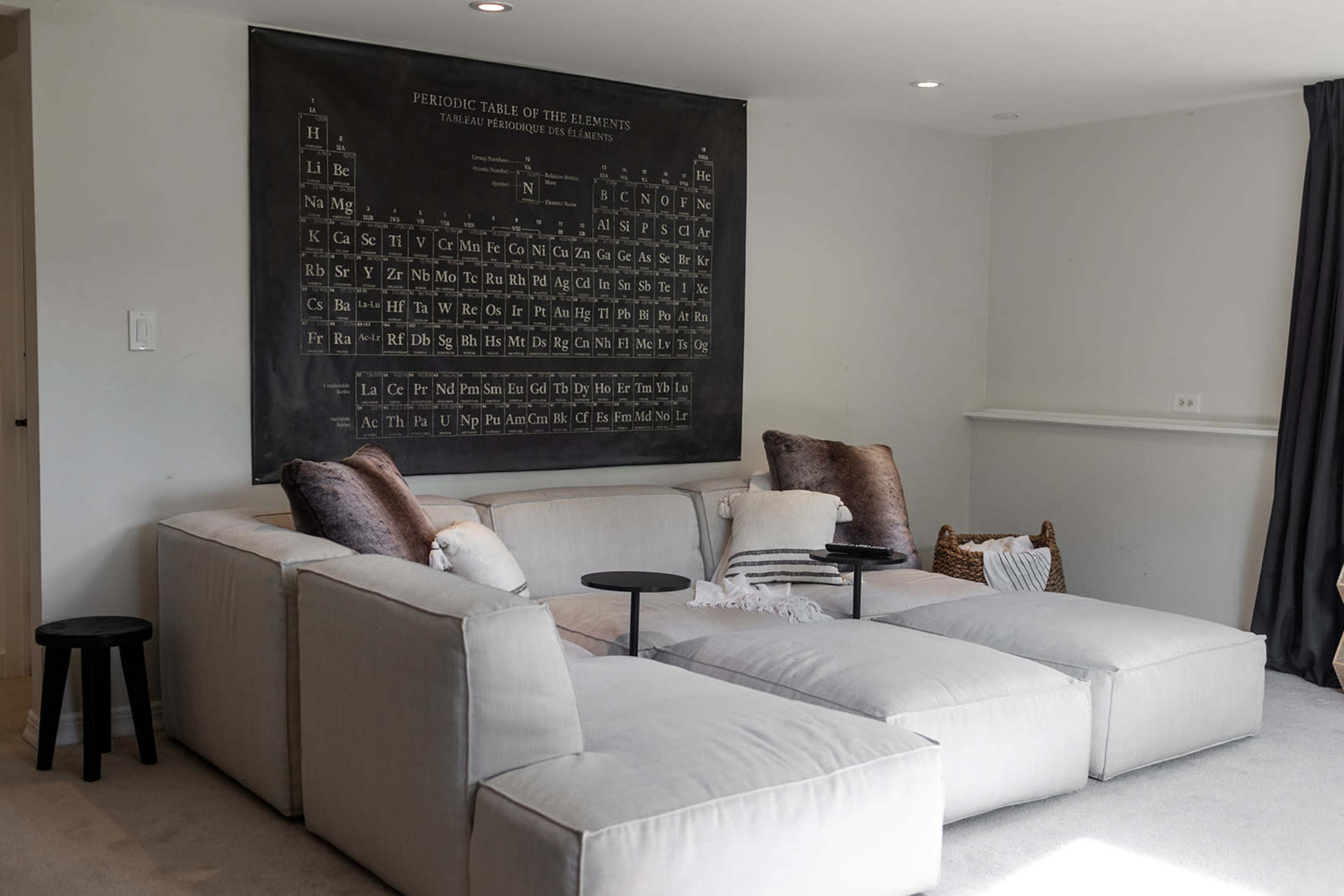 A modern living room features a large sectional sofa with decorative pillows, a small side table, and a chalkboard displaying the periodic table of elements on the wall.