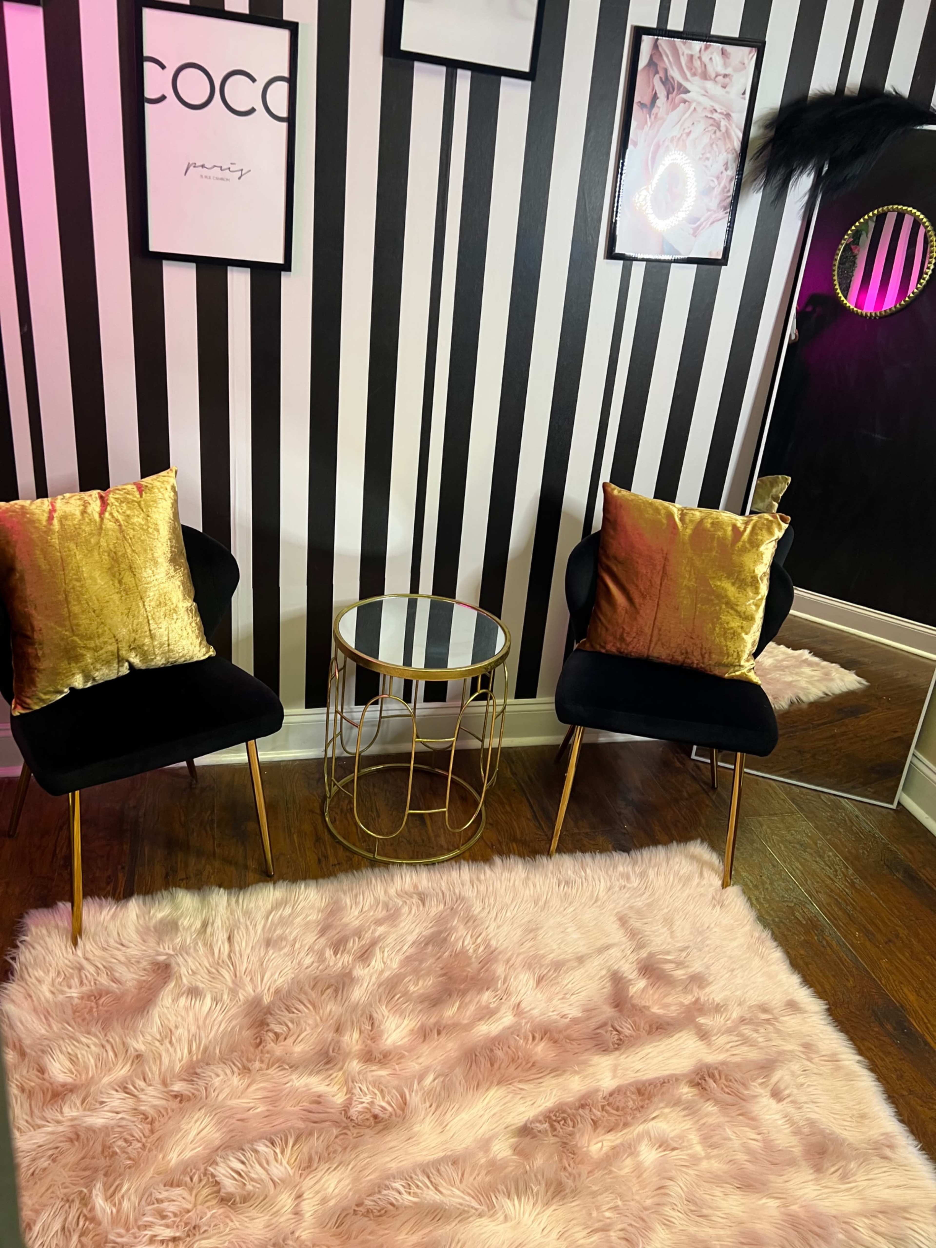 The room features two black chairs with gold accents and decorative pillows, a round glass table, and a plush pink rug against a striped black and white wall.