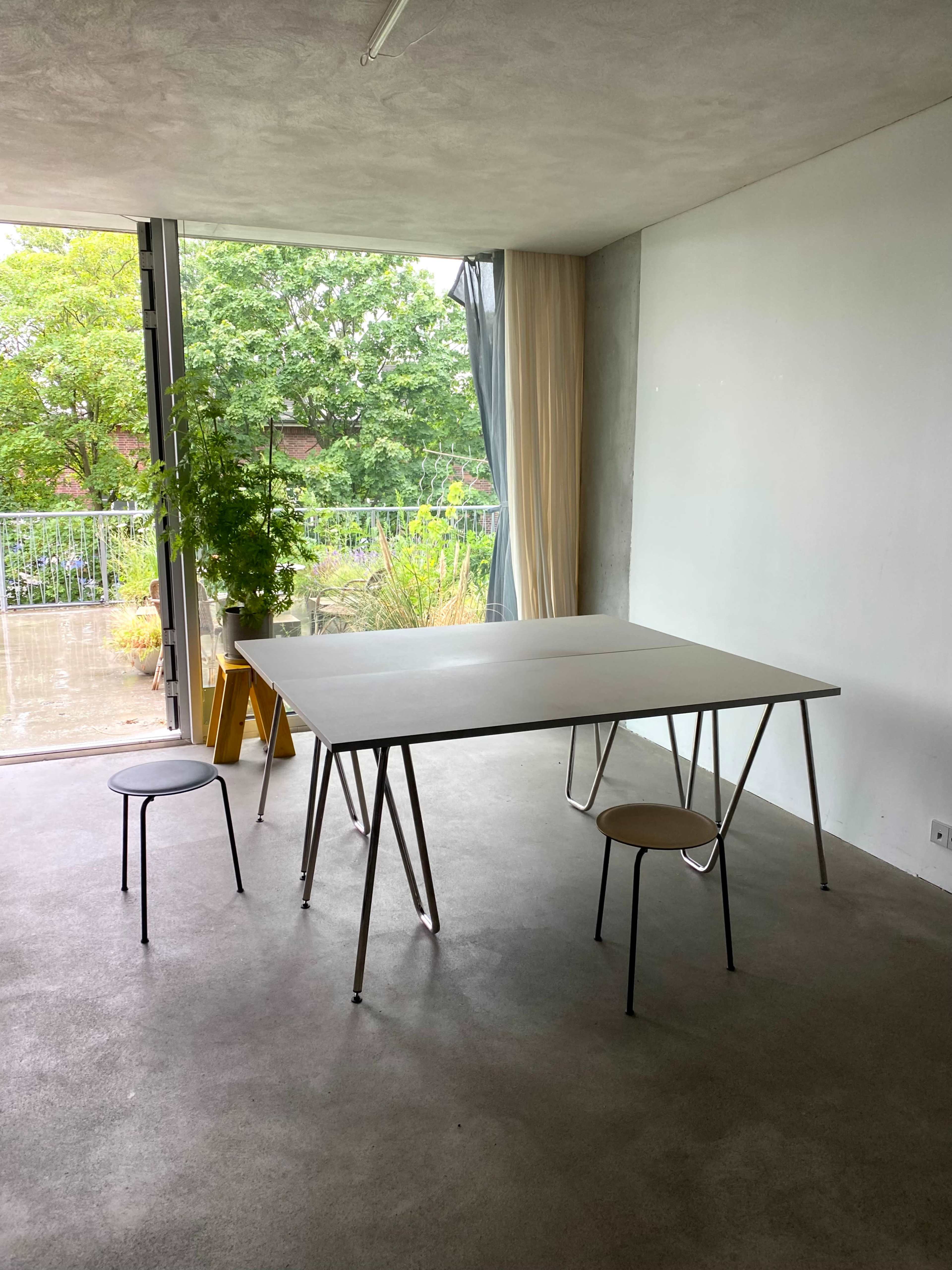 A minimalist room features a large rectangular table with three chairs, surrounded by large windows that show greenery outside.