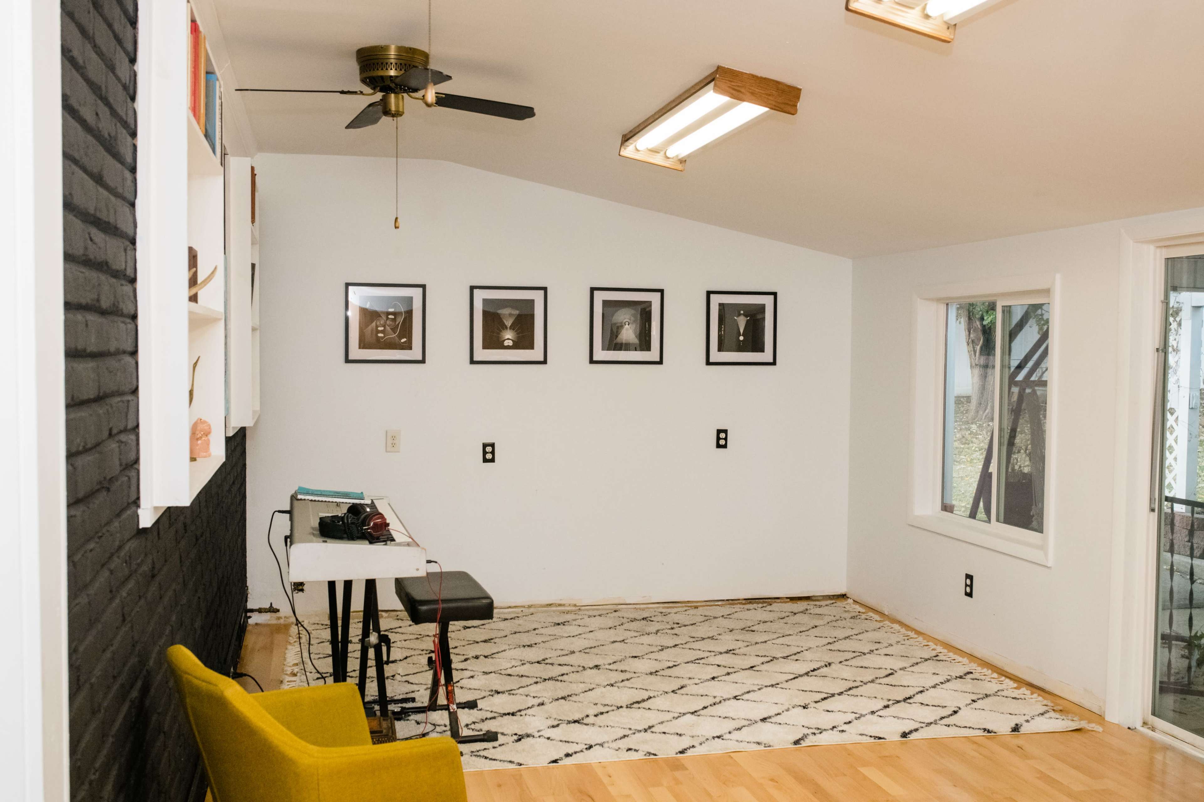 The image shows a sparsely furnished room with a yellow chair, a small table, and black-and-white framed photographs on the wall.