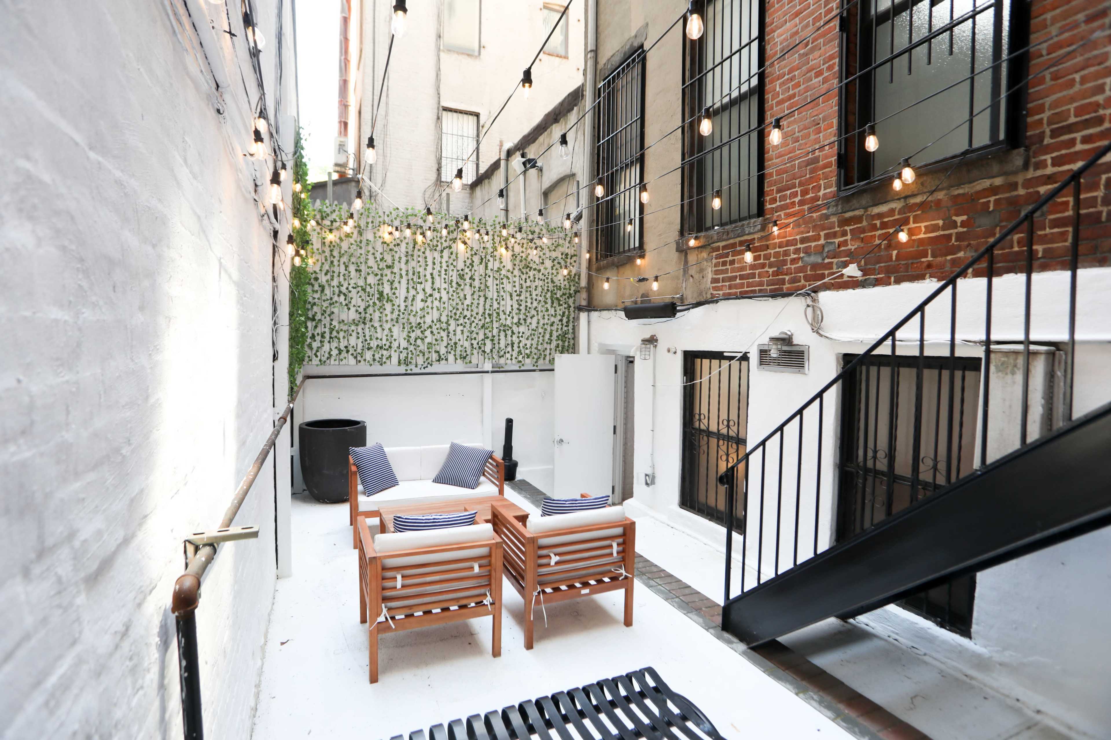 A small outdoor patio area featuring two wooden chairs with cushions, surrounded by white walls, string lights, and a staircase leading to a higher level.