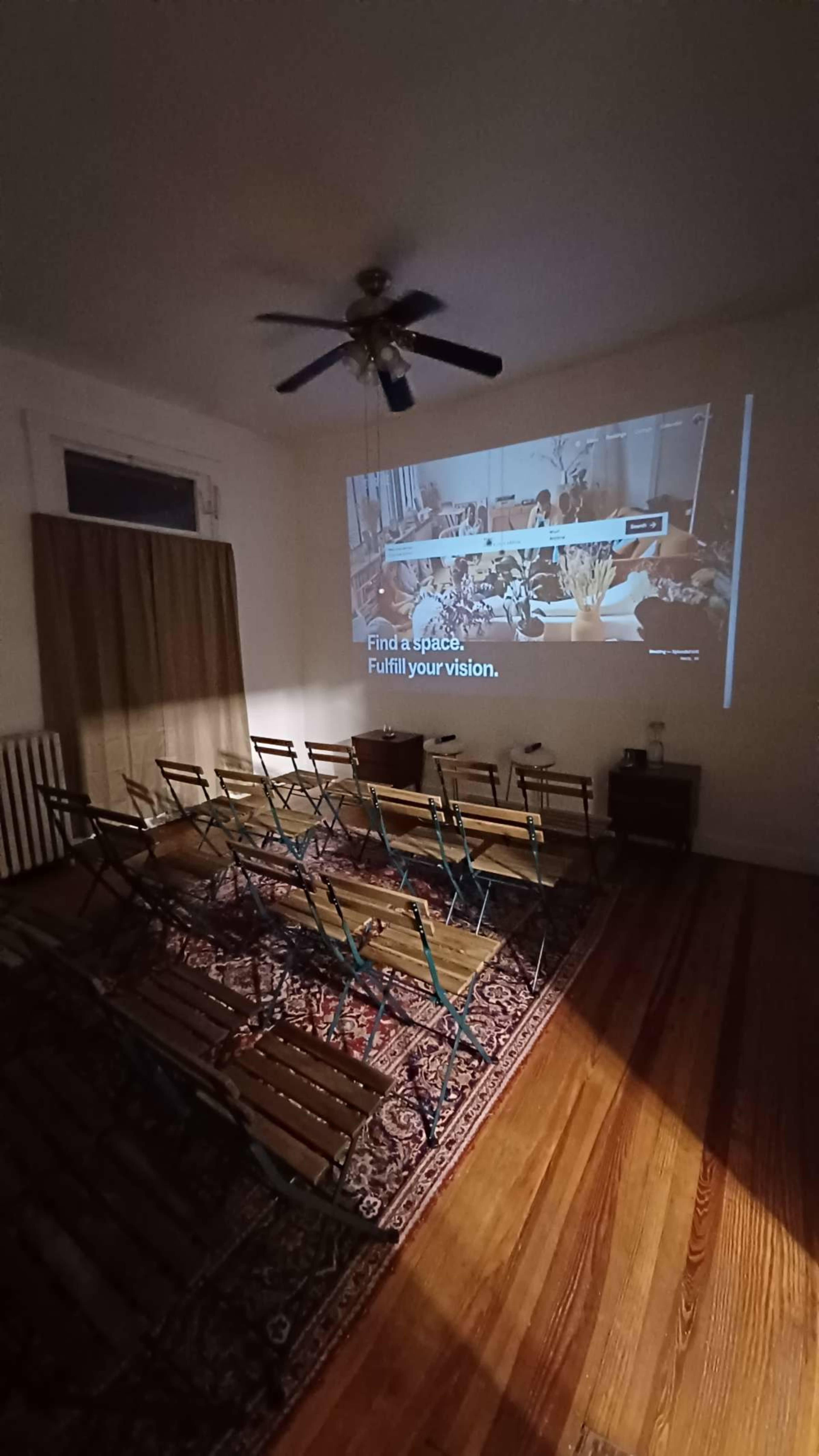 A projector displays a promotional message on a wall in a room with wooden flooring and folding chairs arranged in rows.