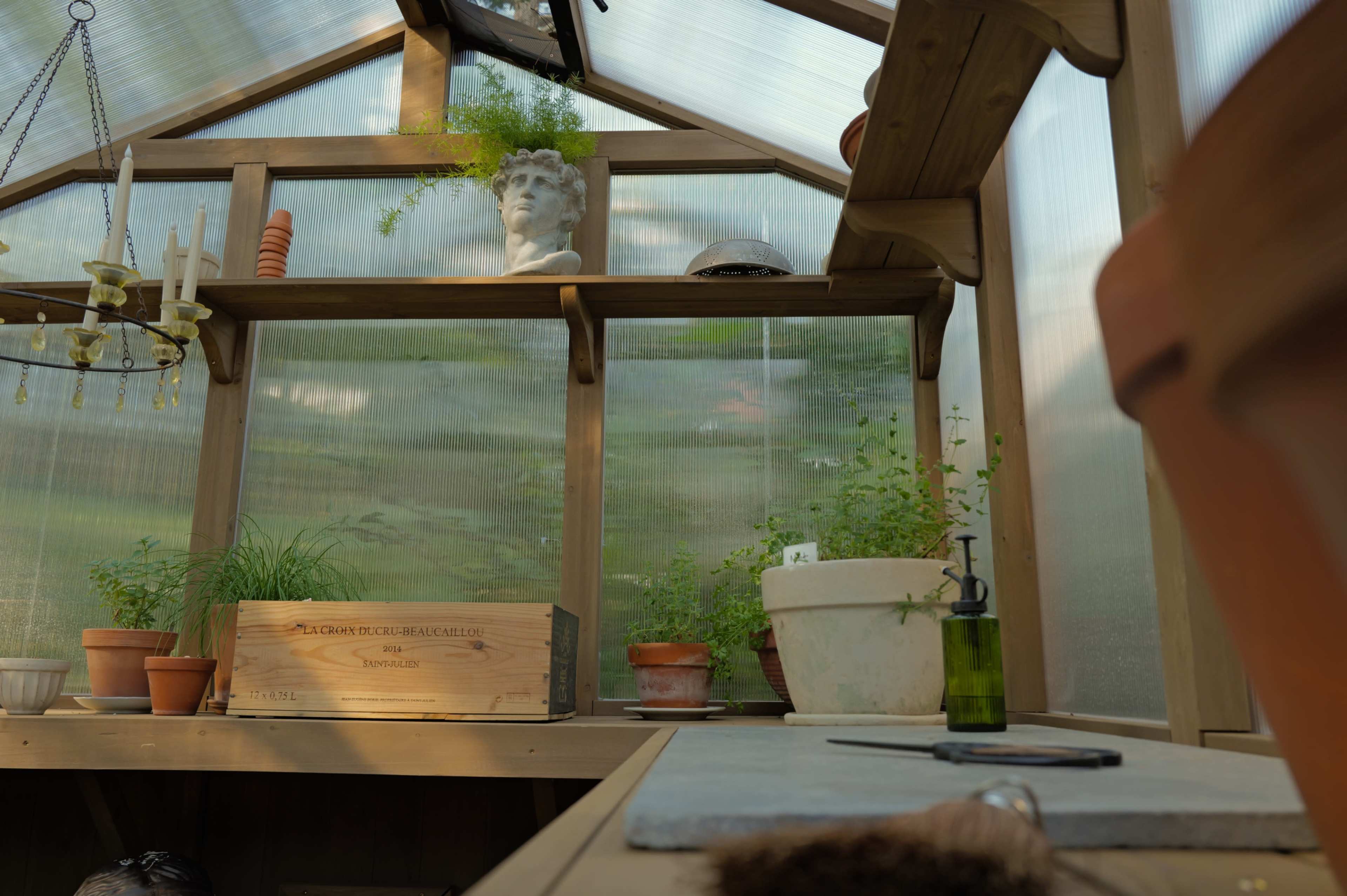 The image shows a wooden greenhouse interior with various potted plants, a stone sculpture, and gardening tools on a table.