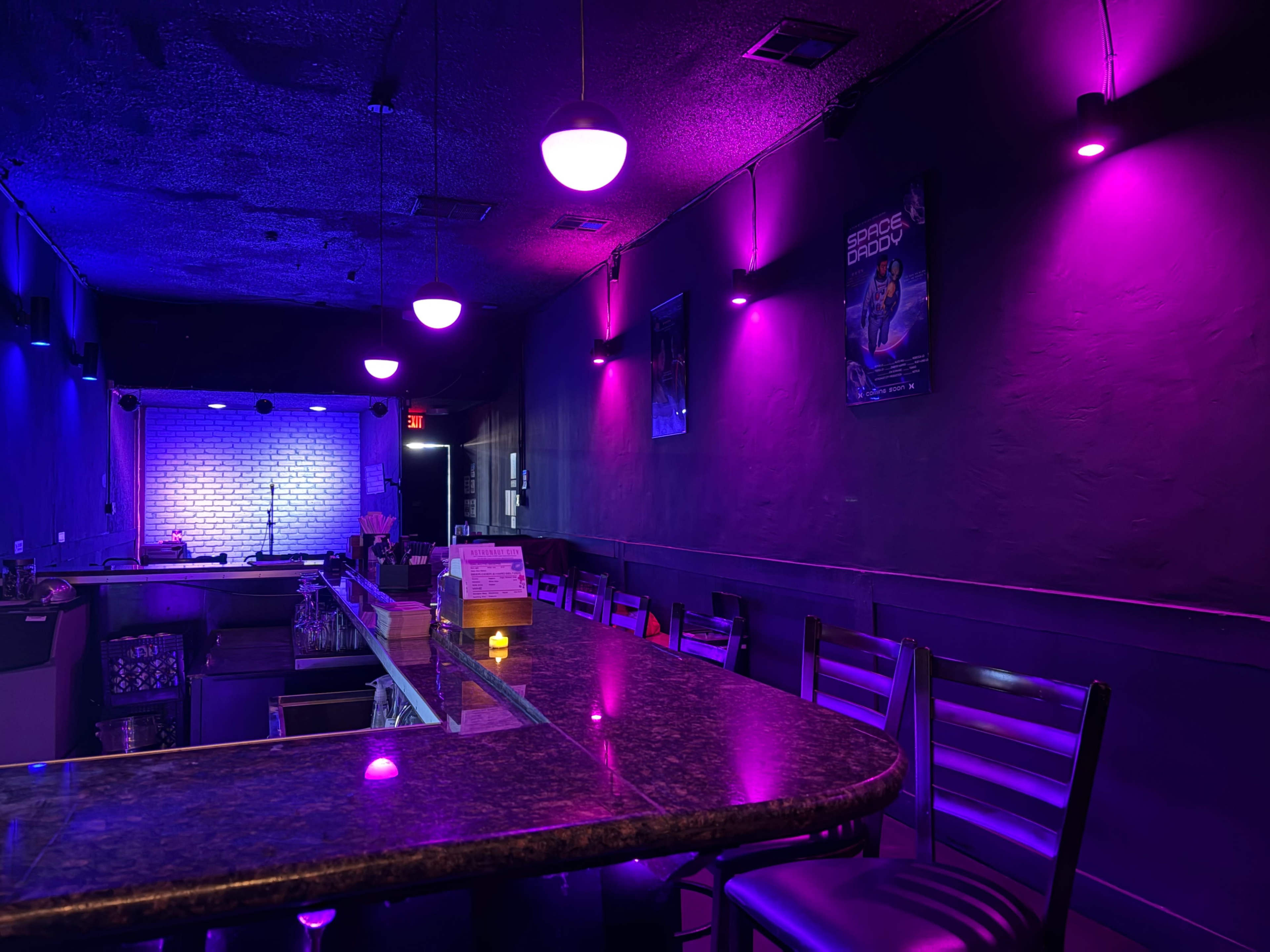 The image shows a dimly lit bar with purple and blue lighting, featuring a long countertop and a performance area in the background.