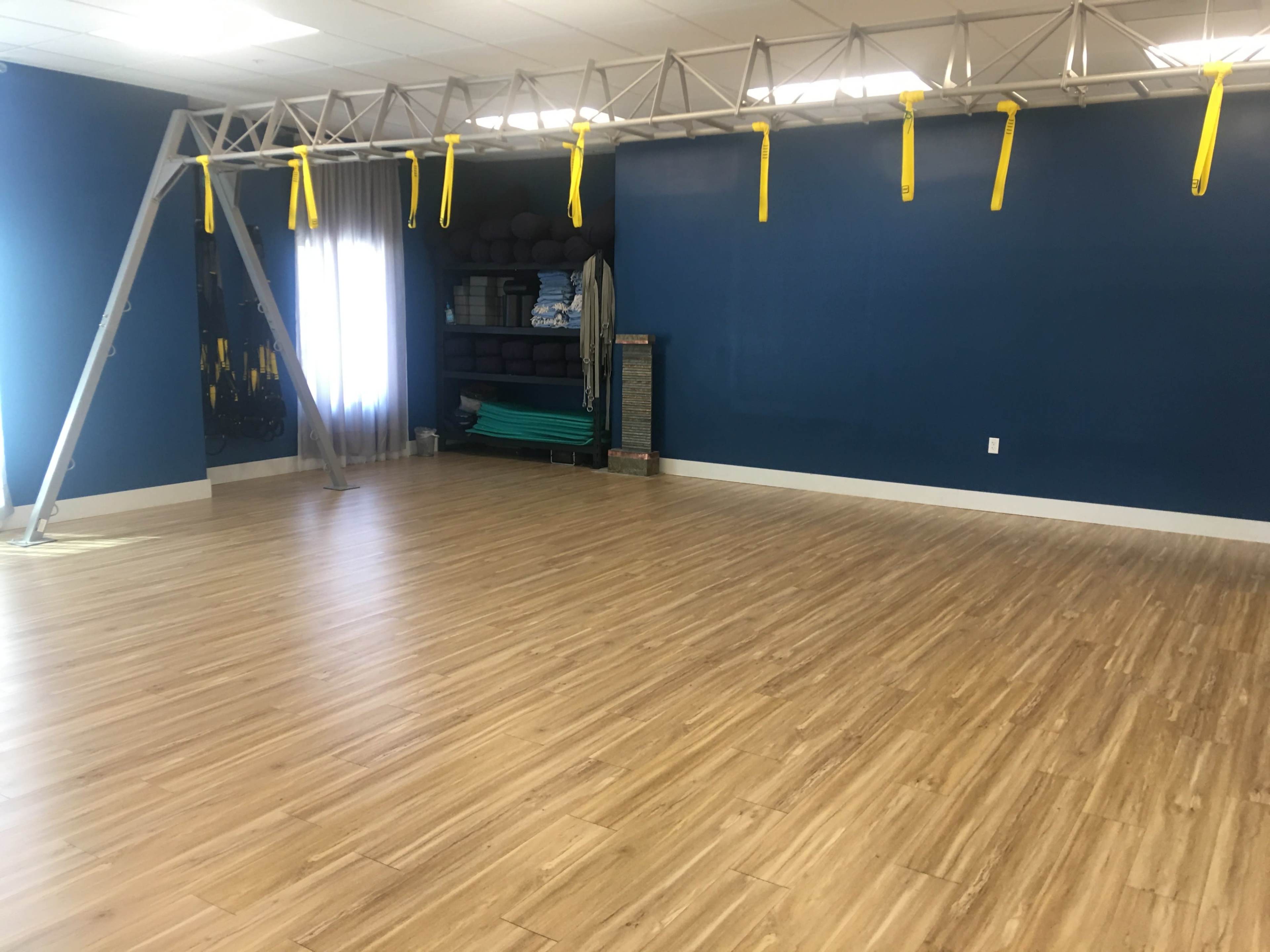 An empty exercise studio with wooden flooring, a yellow-strapped suspension training system suspended from the ceiling, and storage shelves against a blue wall.