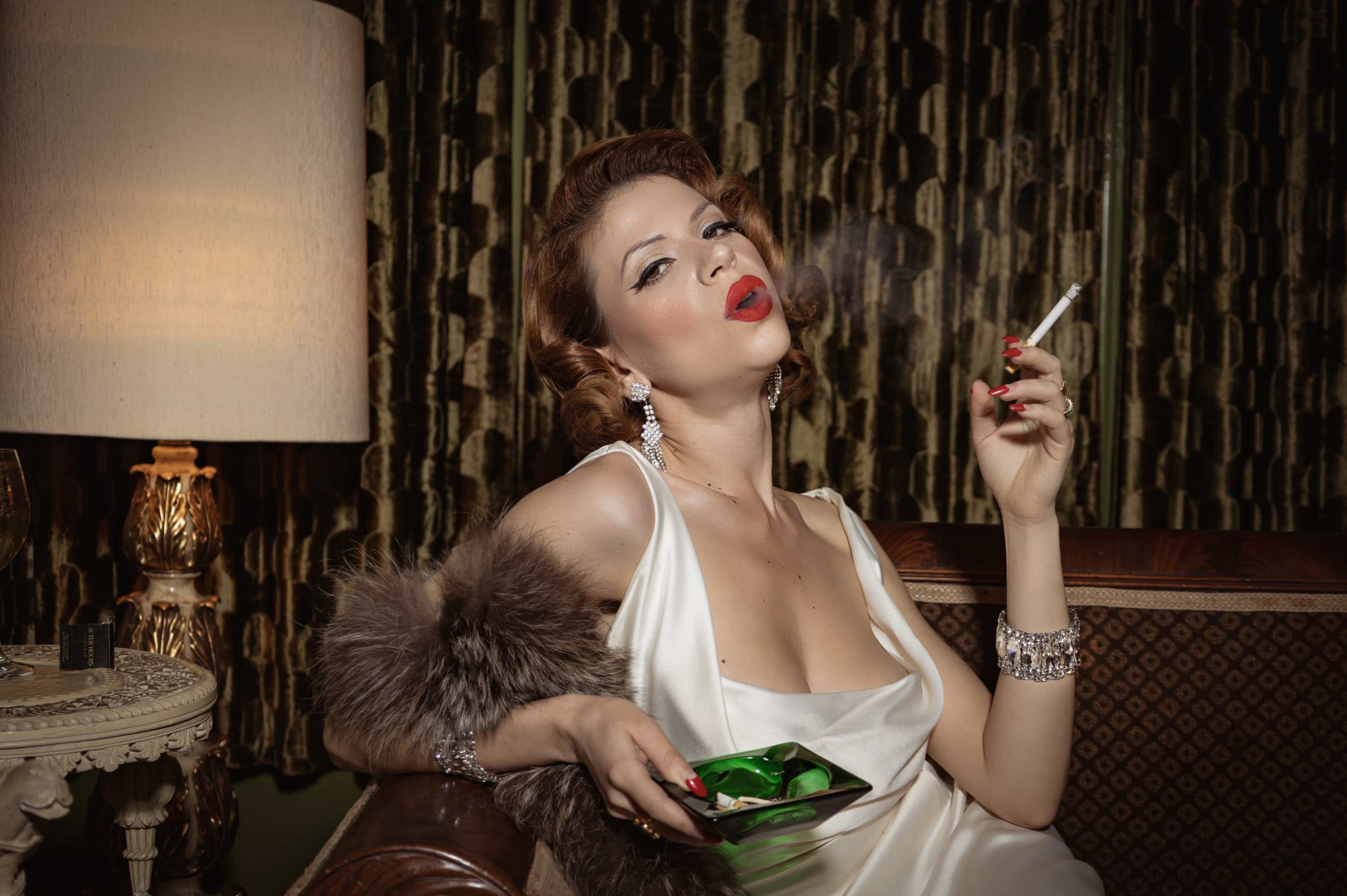 A woman wearing a satin gown sits on a vintage couch, holding a plate and smoking a cigarette in a dimly lit room.