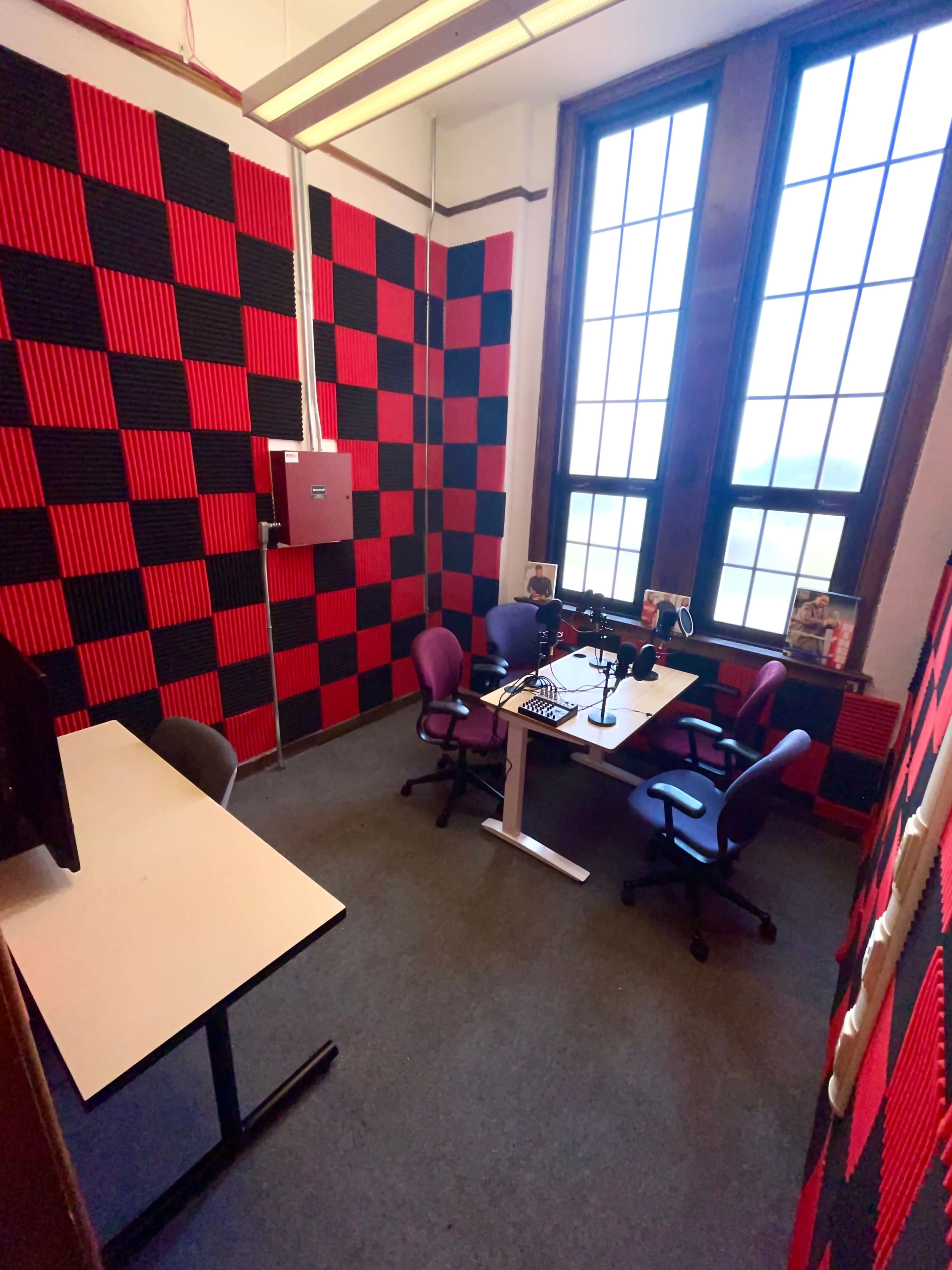 A small, soundproofed room with black and red checkered walls, two chairs, a desk, and equipment for recording or broadcasting.